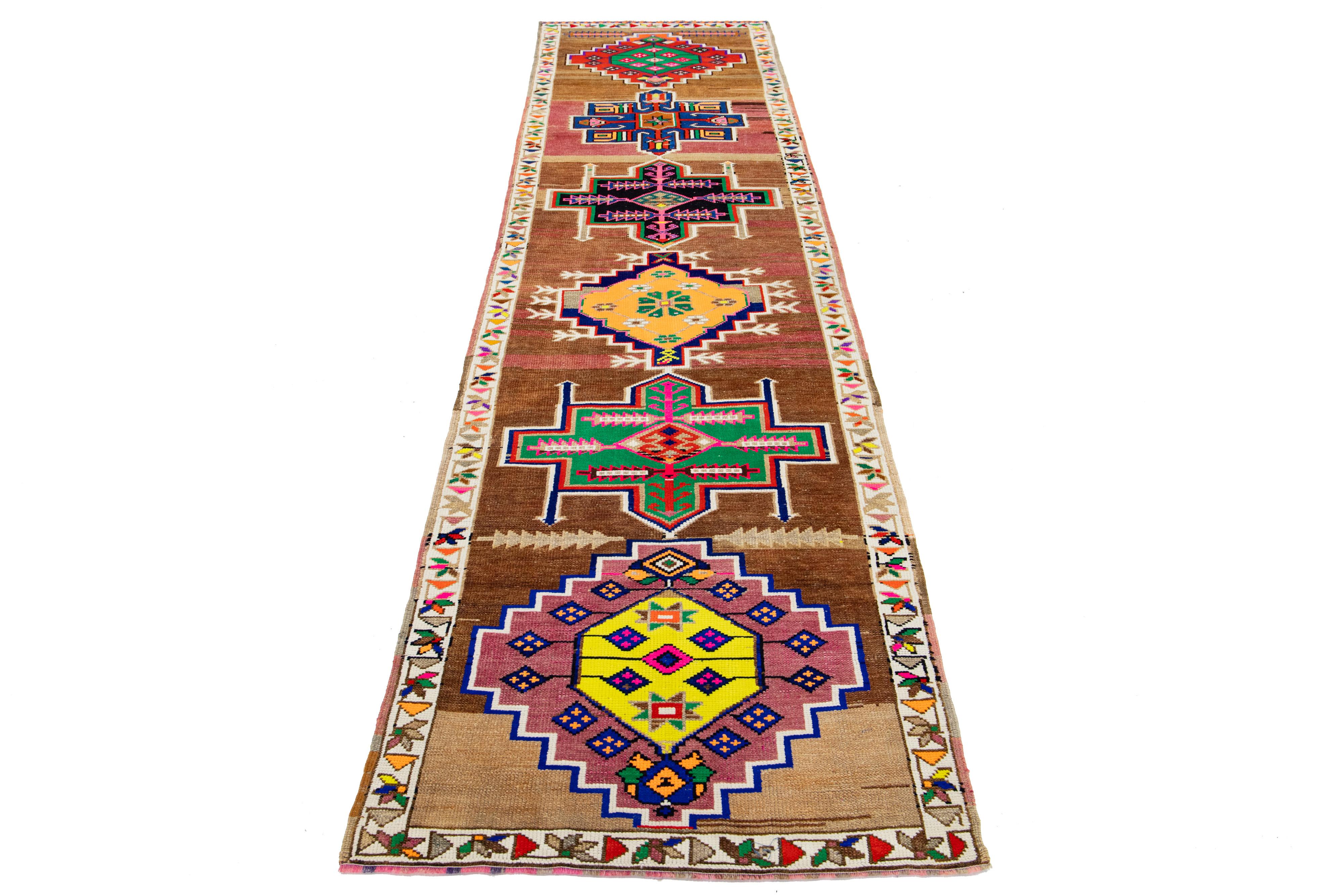 Dieser exquisite Vintage-Läufer wurde in sorgfältiger Handarbeit aus hochwertiger Wolle geknüpft. Es zeichnet sich durch ein sattes braunes Feld aus, das durch mehrfarbige geometrische Akzente aufgewertet wird und sowohl handwerkliches Können als