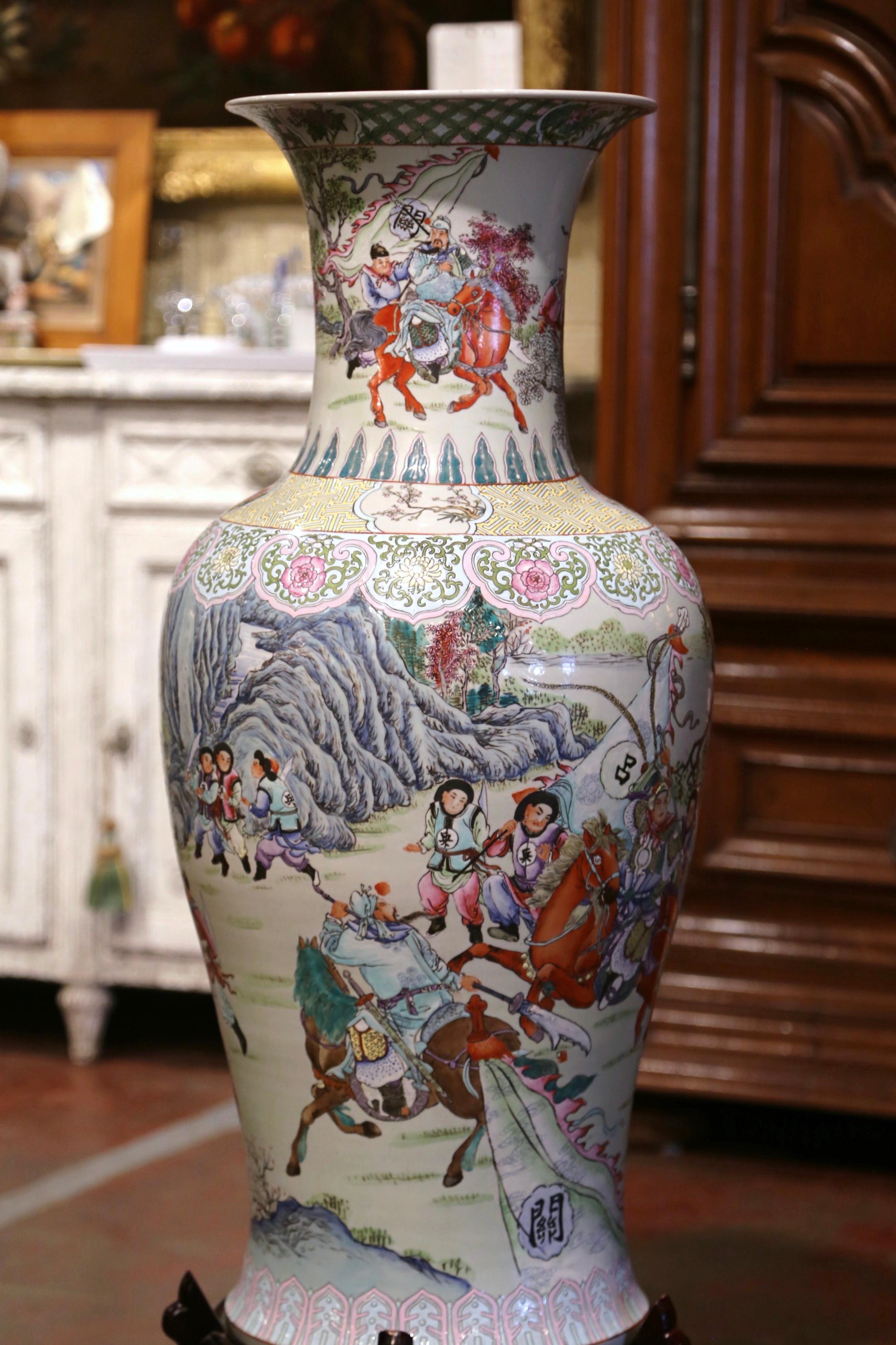 Crafted in China circa 1950, and standing on a carved wooden base, the tall porcelain vase is round in shape with an elegant long and wide neck. The colorful antique urn is decorated with hand painted figural motifs with people on horses. The large