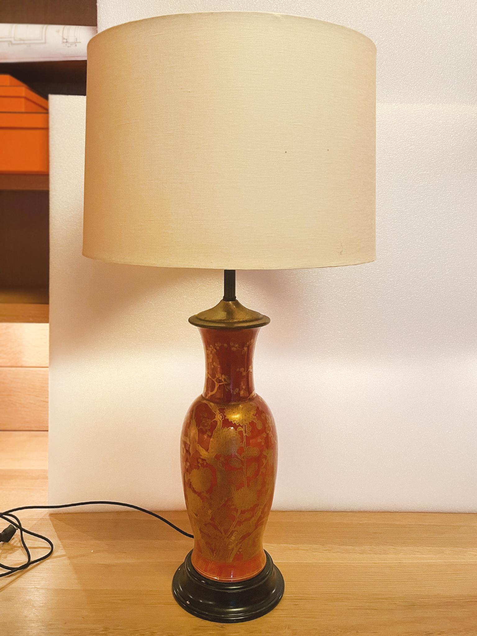 Cette lampe de table chinoise du milieu du 20e siècle se compose d'un corps en métal en forme d'urne, de ferrures en laiton et d'une base en bois ébonisé. Le corps est émaillé d'un riche rouge décoré de motifs de fleurs et d'oiseaux en or. La lampe
