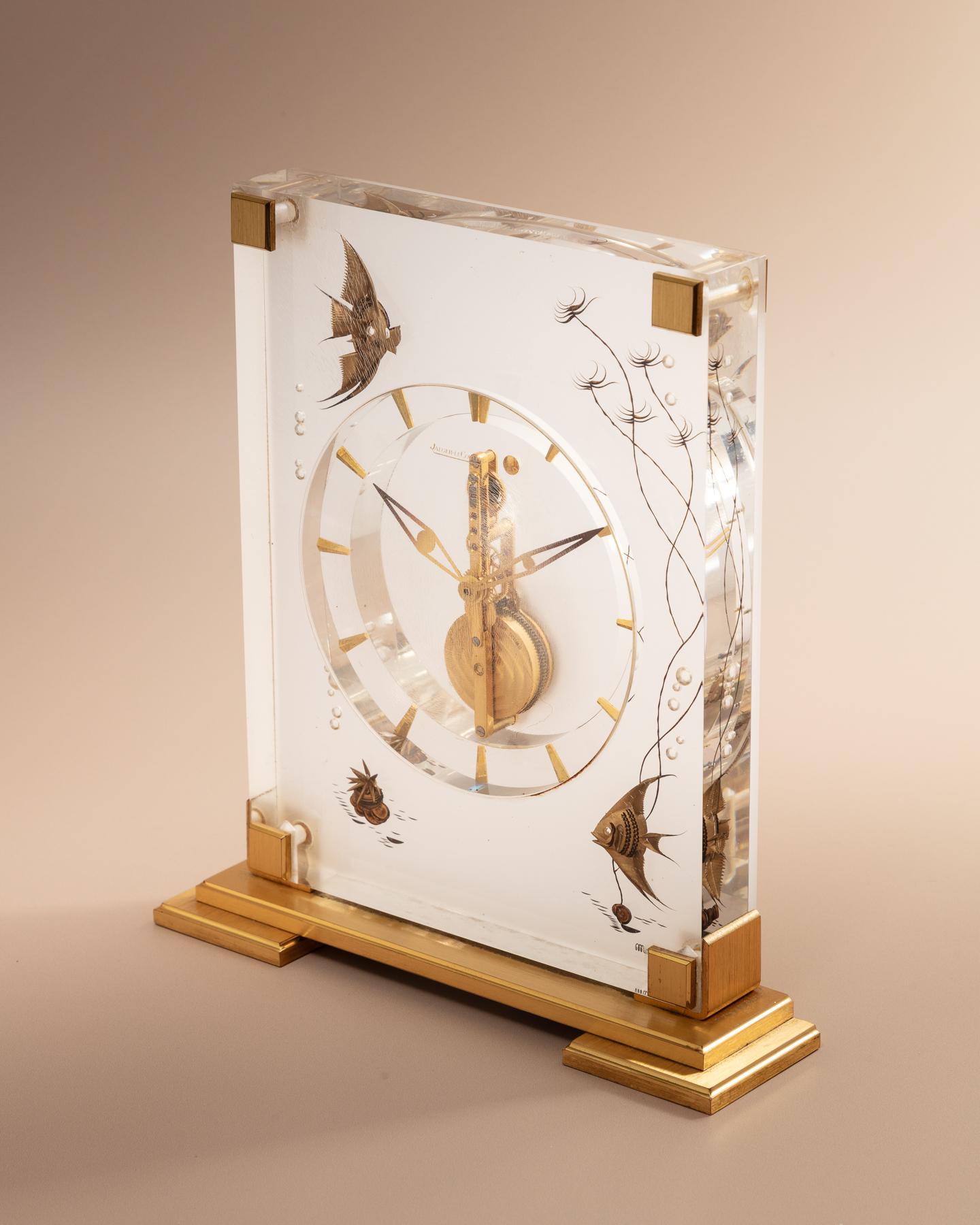 A mid-century mantel or desk clock by Jaeger-LeCoultre, set within an elegant lucite case decorated with inset brass motifs of fish and rising air bubbles. 
The clock runs on an eight-day movement and features Jaeger-LeCoultre’s signature numerals