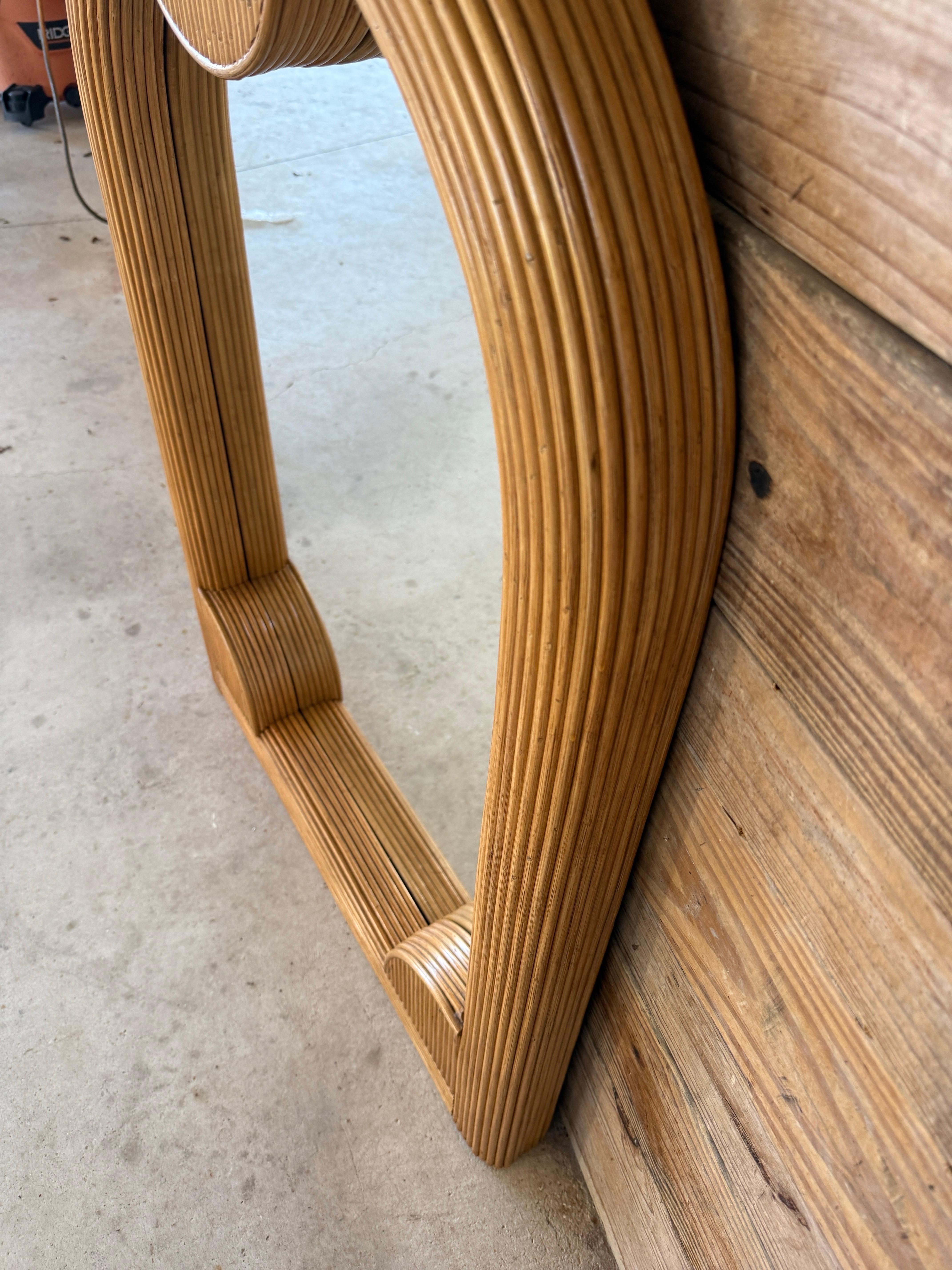 triking Mid-20th Century pencil rattan wall mirror featuring a sculptural arched design with geometric half-moon accents. Fully refinished, showcasing the natural honey tones and ribbed texture of the rattan. A new mirror has been added, making it