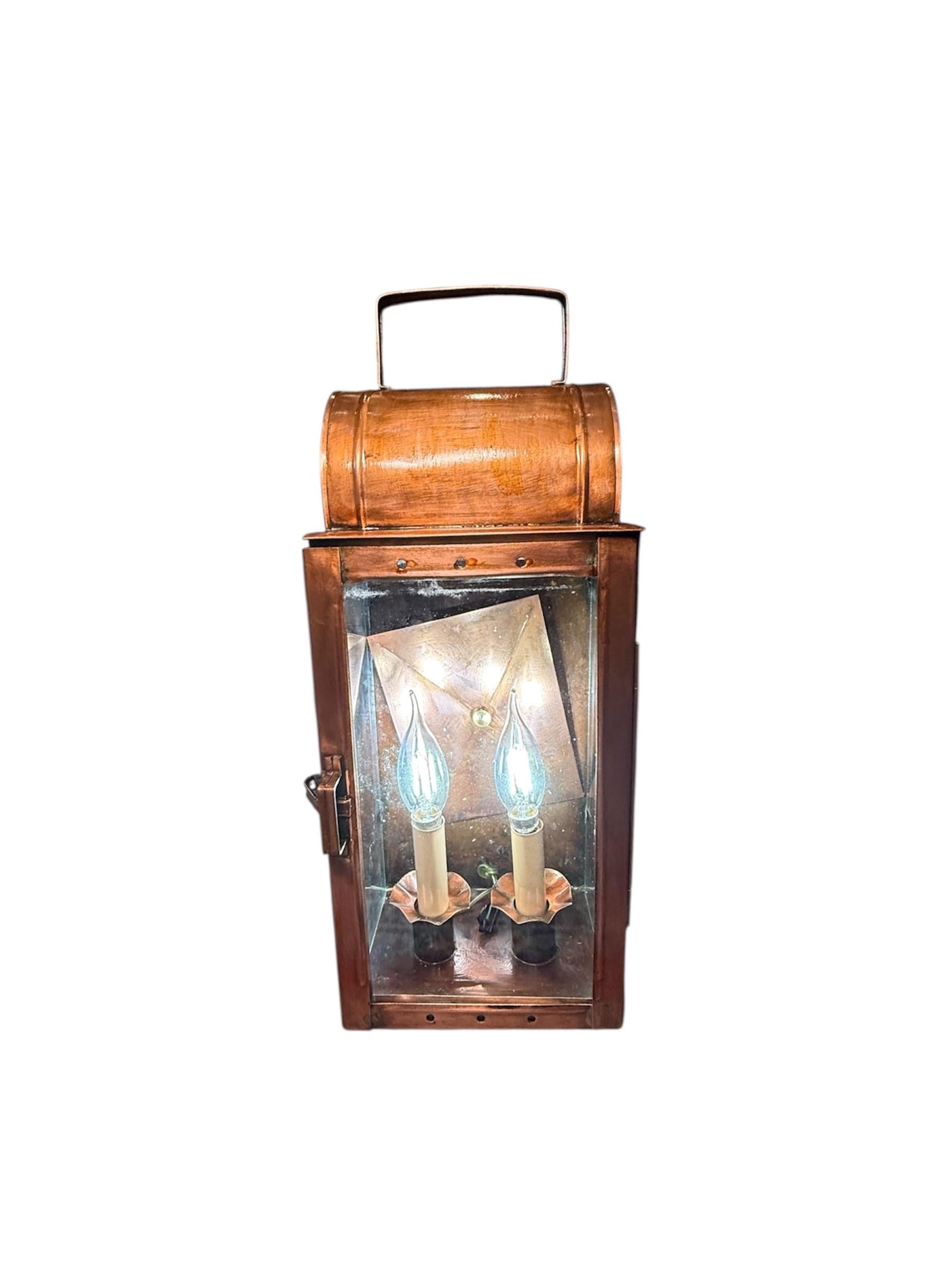 Este es un farol de pared de estilo colonial de mediados del siglo XX electrificado. Representa una caja rectangular de latón y cristal, de color cobre, con techo abovedado y asa adosada. Hay dos velas como bombillas eléctricas dentro de la caja de