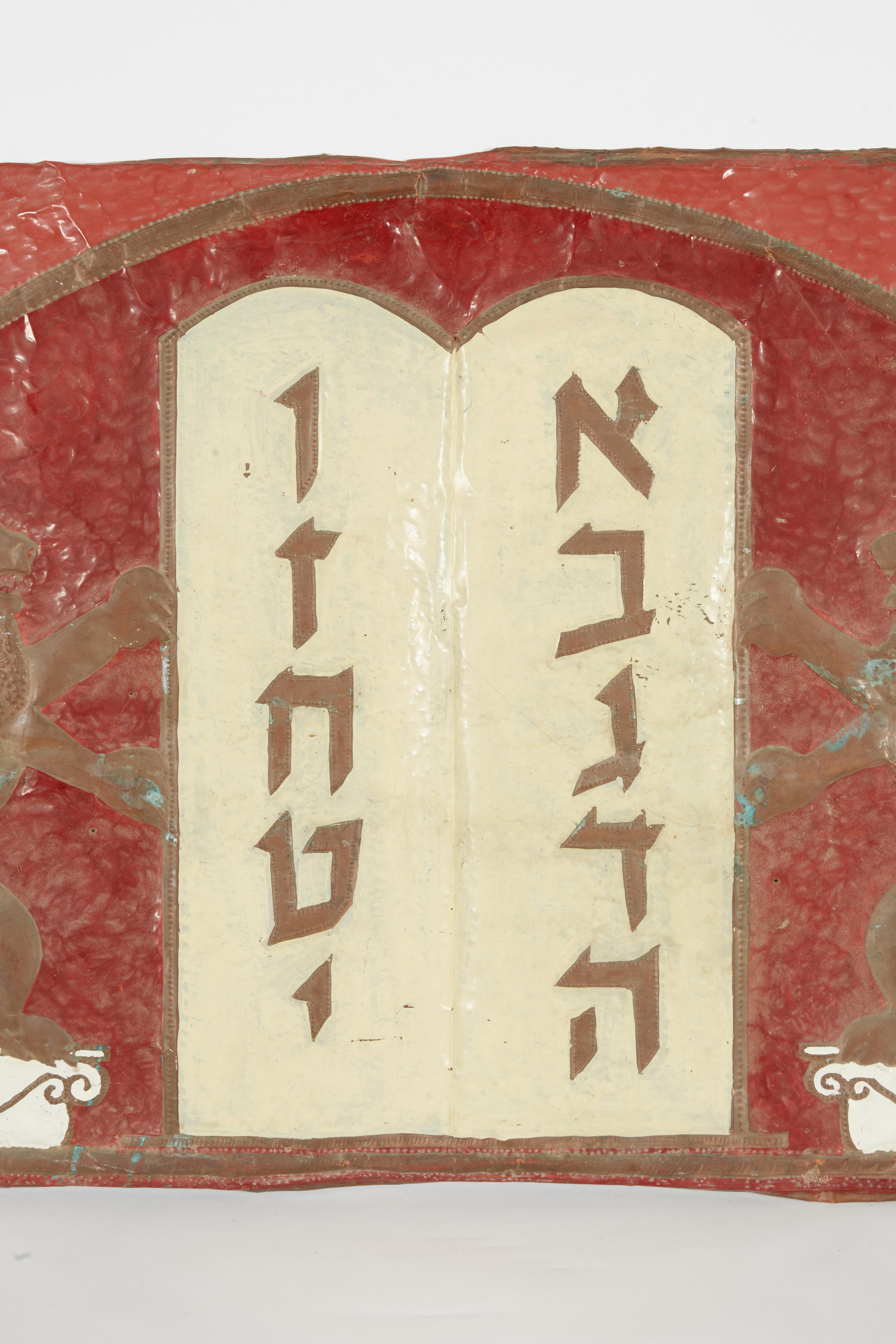 Hand hammered copper sheet synagogue decoration depicts the Decalogue (Ten Commandments) flanked by rampant lions standing on a column, framed by an arch with two stars of David at the top corners.

 