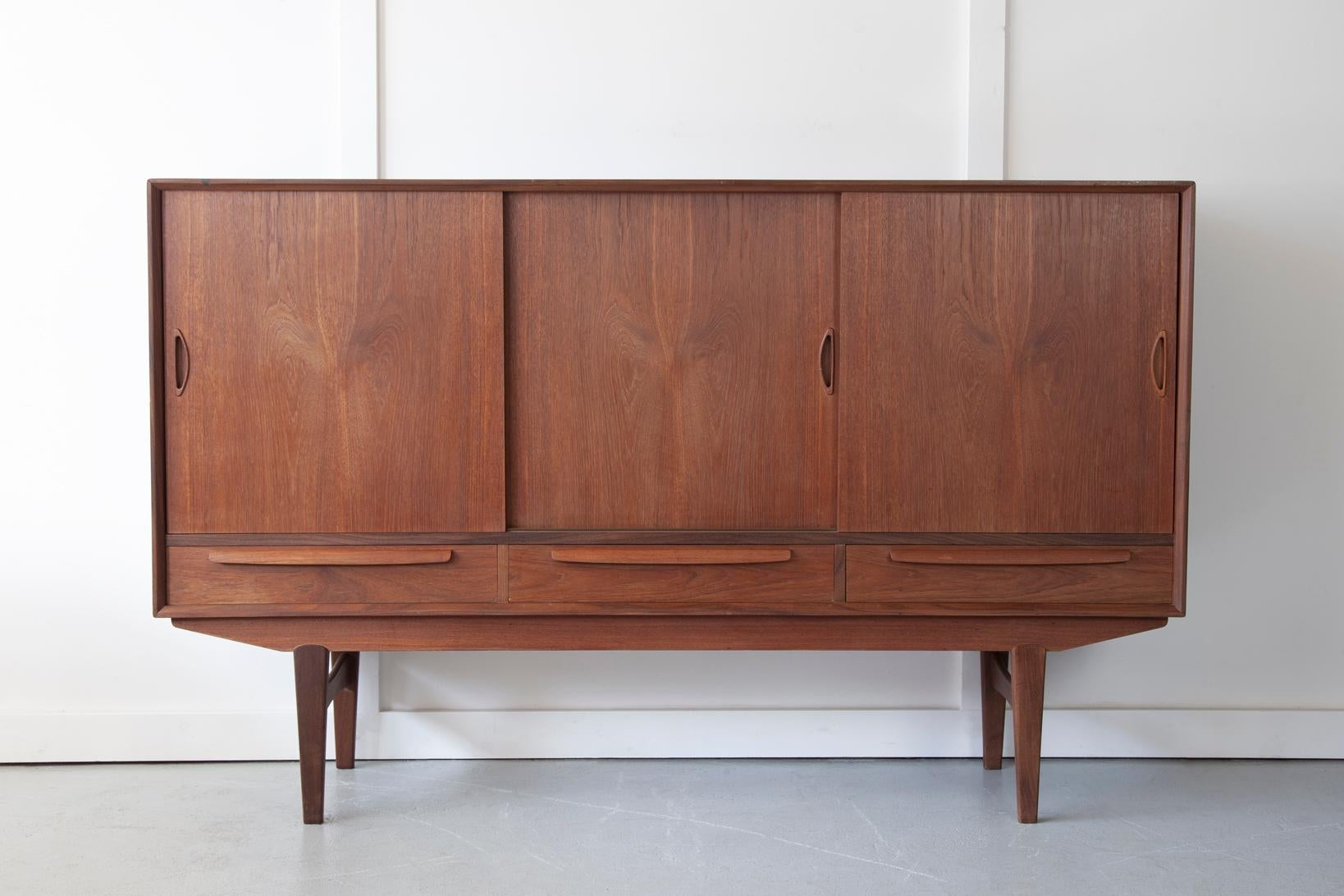 A large teak highboard from Denmark, with three drawers which feature elegant lipped handles and sliding doors with curved inset handles to reveal expansive storage compartments. The middle compartment houses a set of three shallow drawers and a