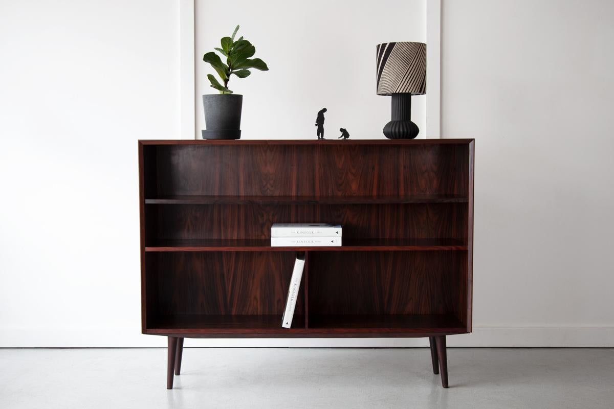An elegant rosewood bookcase, made in Denmark in the 1960's. The top shelf is adjustable allowing it to be moved up and down to suit different sized books and objects. 

This bookcase a small square cut out in the backboard which was made by a