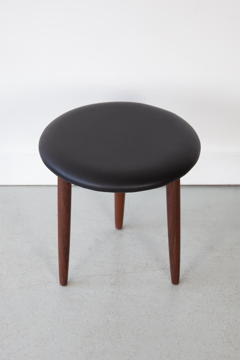 A charming, round footstool with tapering teak legs and a padded cushion which has been newly upholstered in black faux leather. 