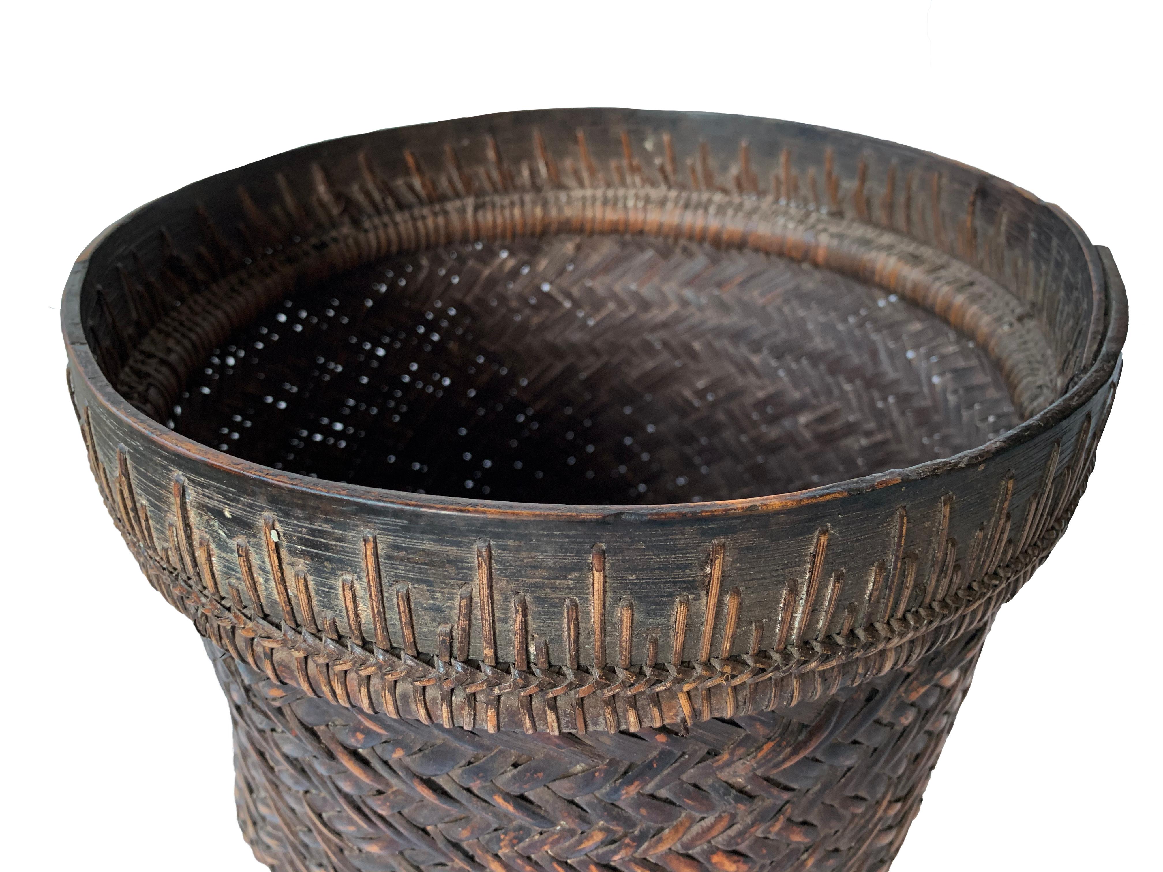 Questo cesto intrecciato a mano della metà del XX secolo proviene dalla tribù Dayak del Borneo ed è realizzato con fibre di rattan e un bordo in legno di ferro. Questi cesti venivano utilizzati per trasportare foglie, frutta, cereali e legno e sono