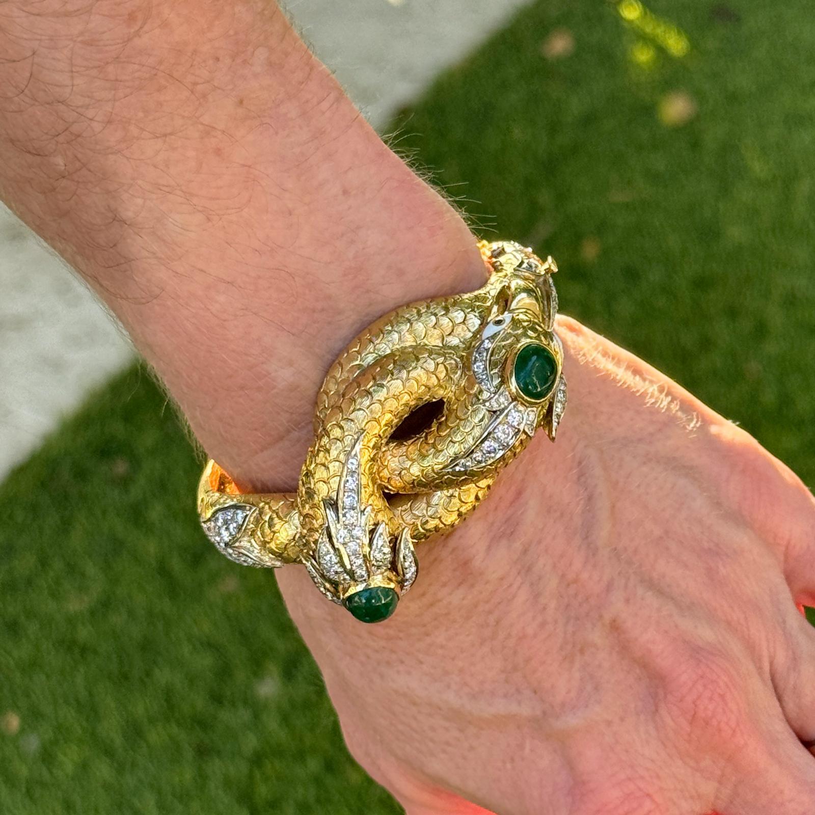 A striking mid-20th-century design, this textured 18K yellow gold hinged bangle features two sculpted snake heads meeting at the center, symbolizing eternity and protection. The heads are set with two cabochon emeralds and accented by approximately