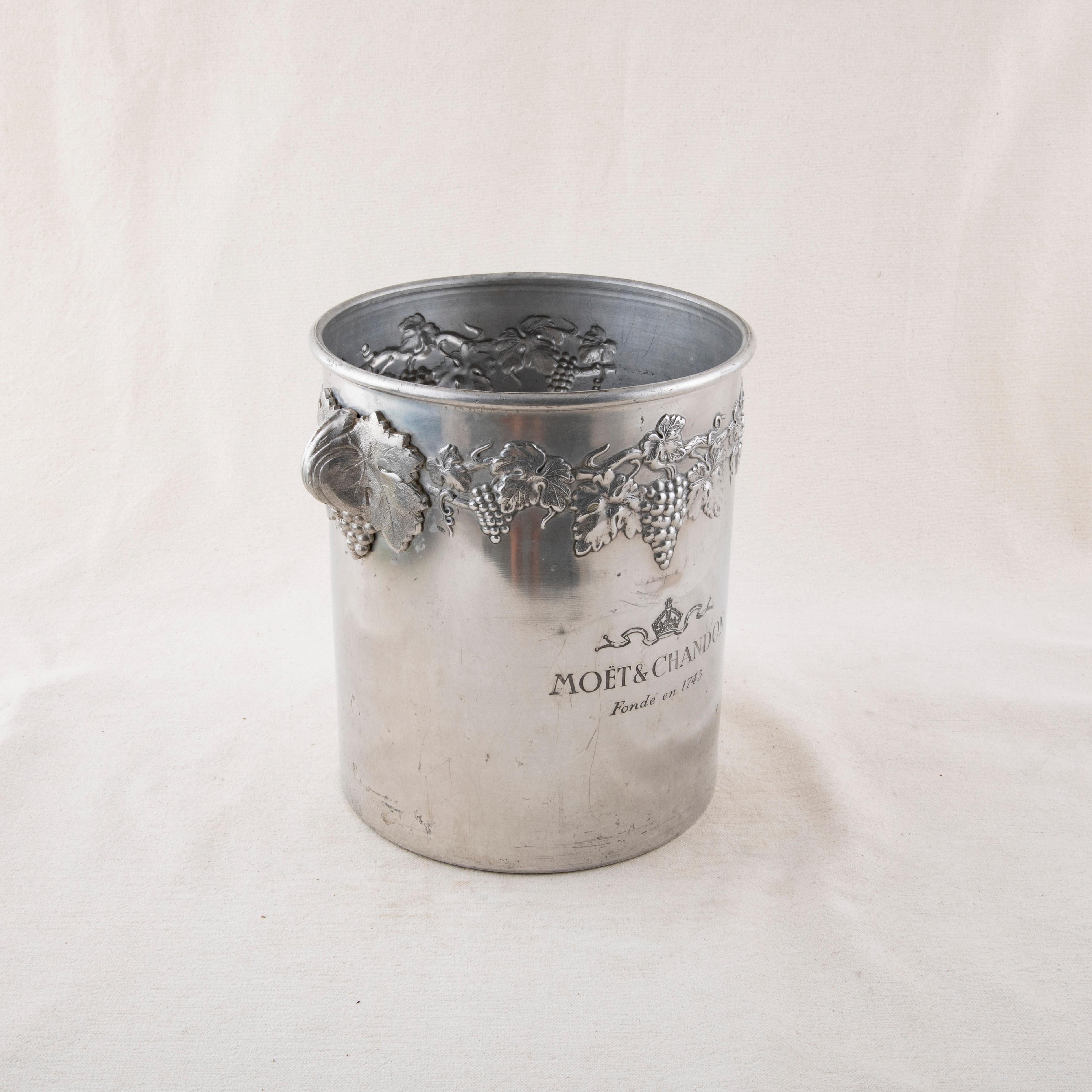 Este cubo de champán francés de aluminio de mediados del siglo XX presenta uvas y hojas de parra alrededor del borde superior. El cubo lleva la marca Moet et Chandon, Fonde en 1743, el nombre del fabricante de champán y el año de fundación de la