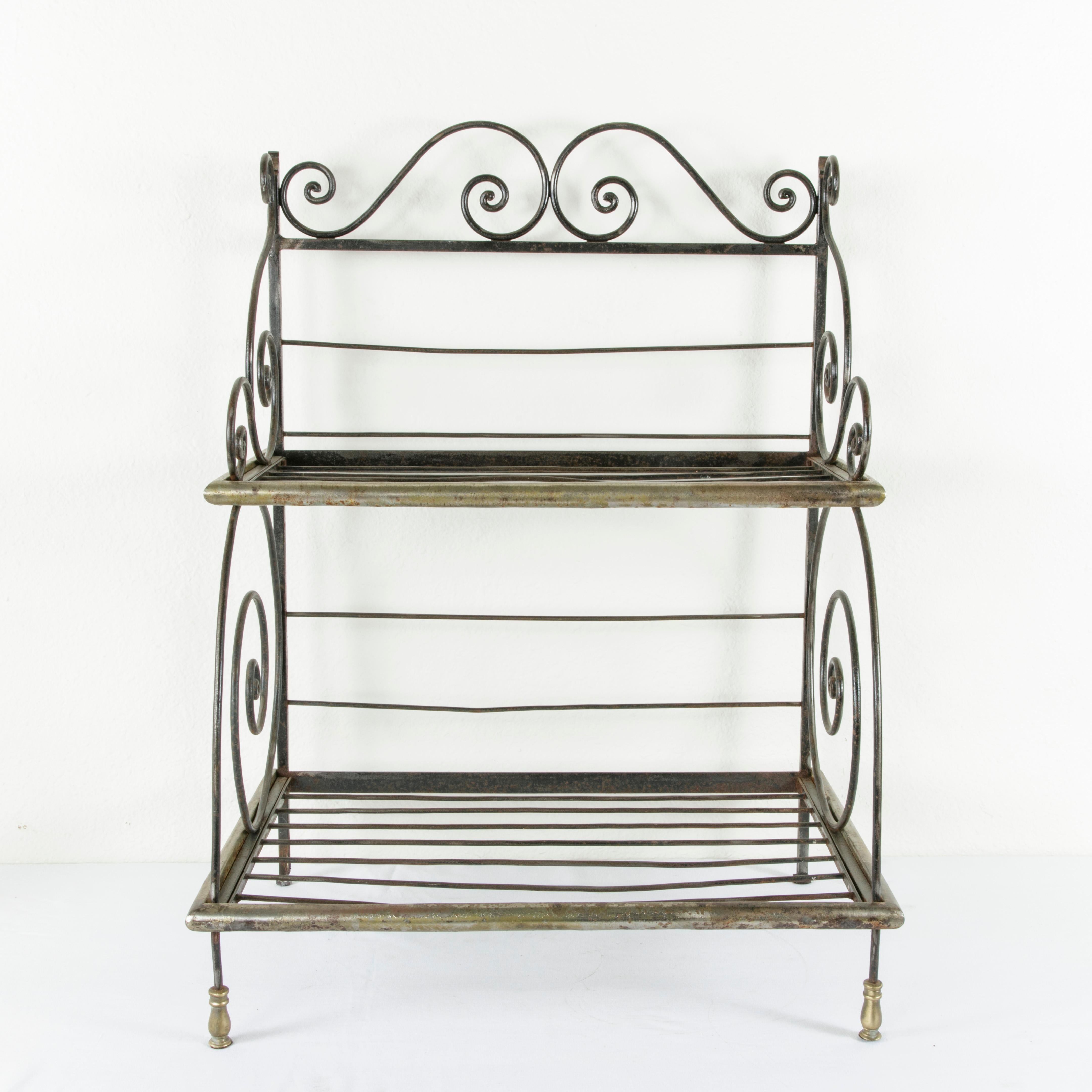 Originally placed upon a counter in a French boulangerie or bakery, this artisan-made iron baker's rack from the mid-20th century features brass detailing on its scrolling form. Brass plated steel banding surrounds the edges of its two shelves. Its