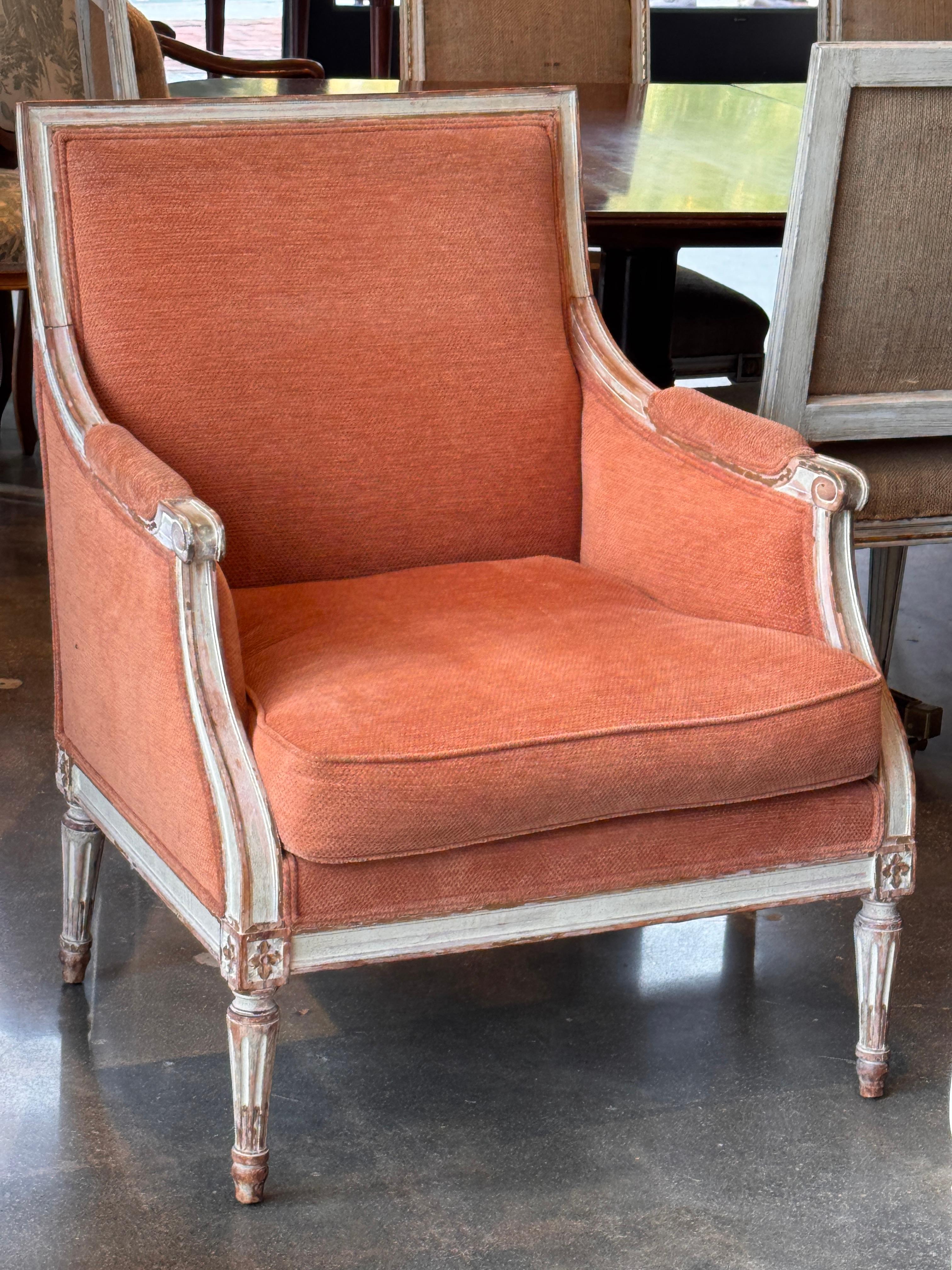 Bergère française à dossier en forme, accoudoirs en volute et pieds cannelés. Le cadre conserve une finition peinte en relief avec des rosettes sculptées dans les coins. Tapissé d'un tissu rouille texturé avec une assise à coussin lâche et des