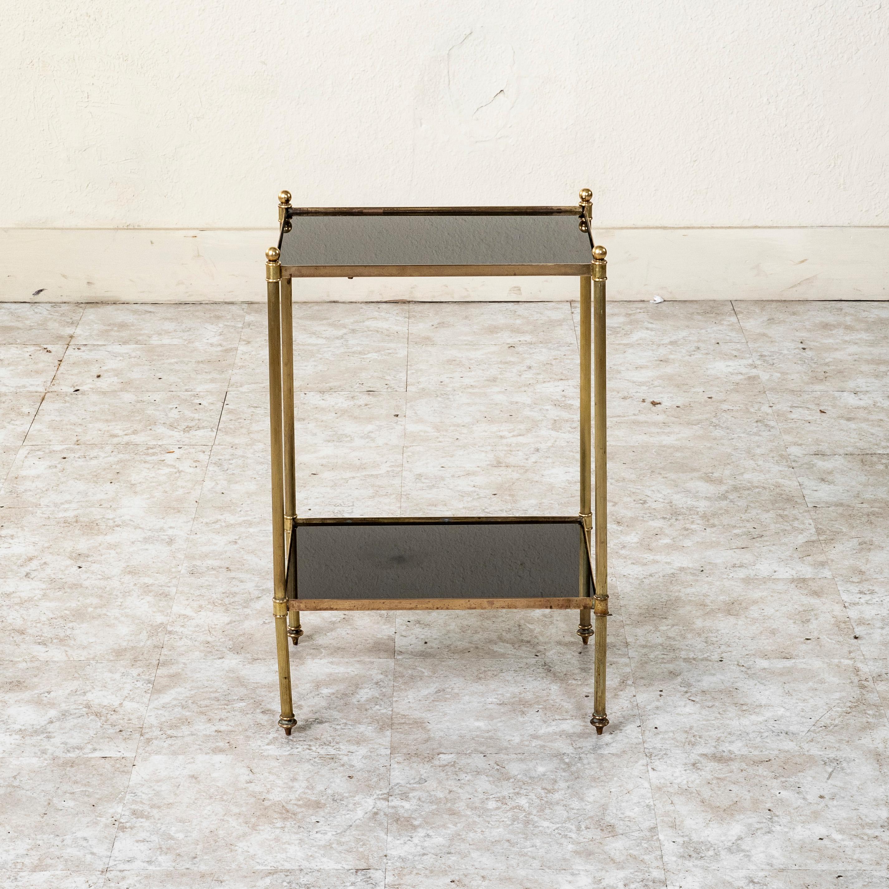 This small scale mid-century French brass side table features an upper and lower shelf fitted with its original black glass. The table rests on fluted legs capped with brass finials and finished with finial feet. c. 1960