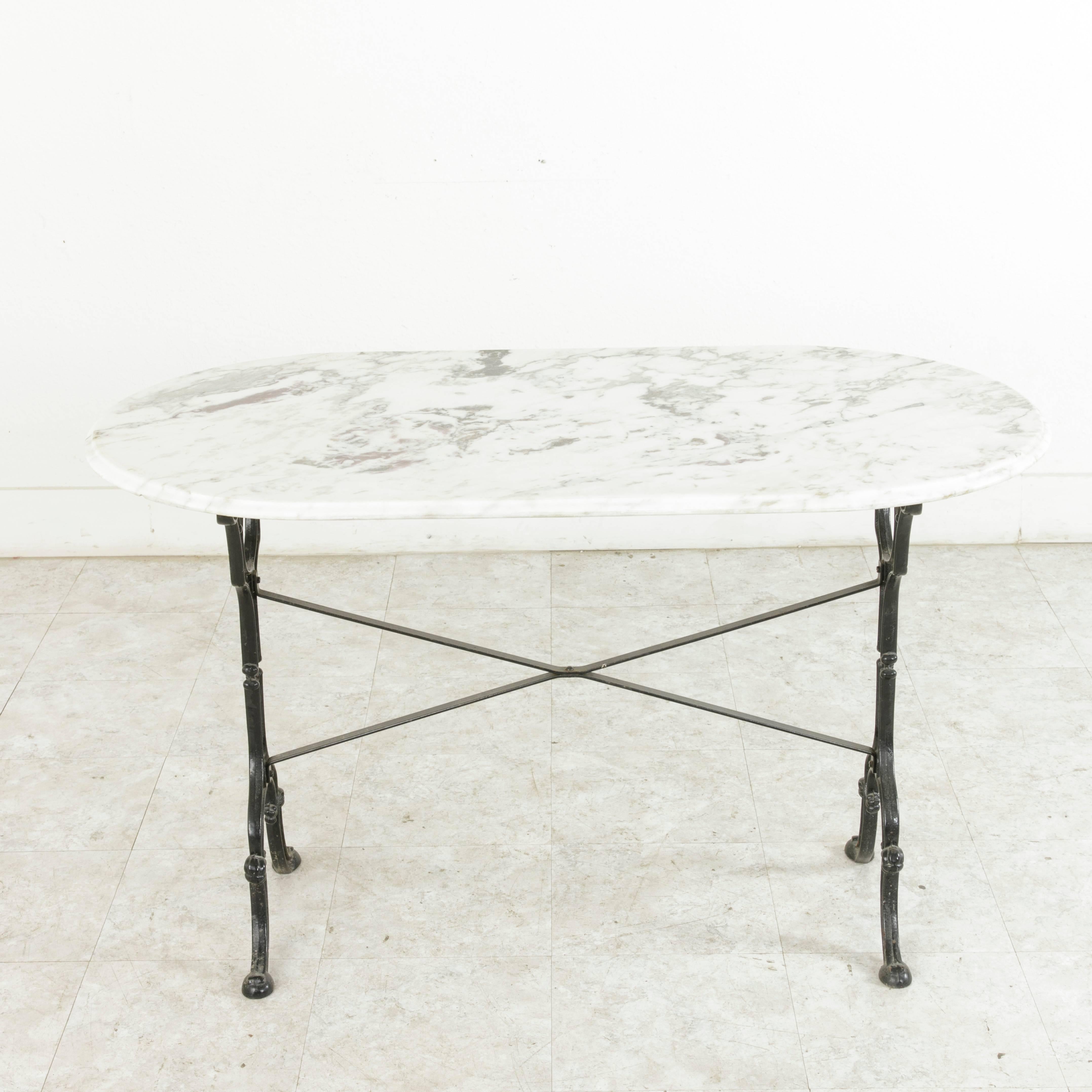 This early 20th century French cast iron bistro table features a bevelled oval white marble top enhanced by purple and grey veining. The curved legs adorned with rosettes and shells are joined by an X-stretcher that provides extra stability to the