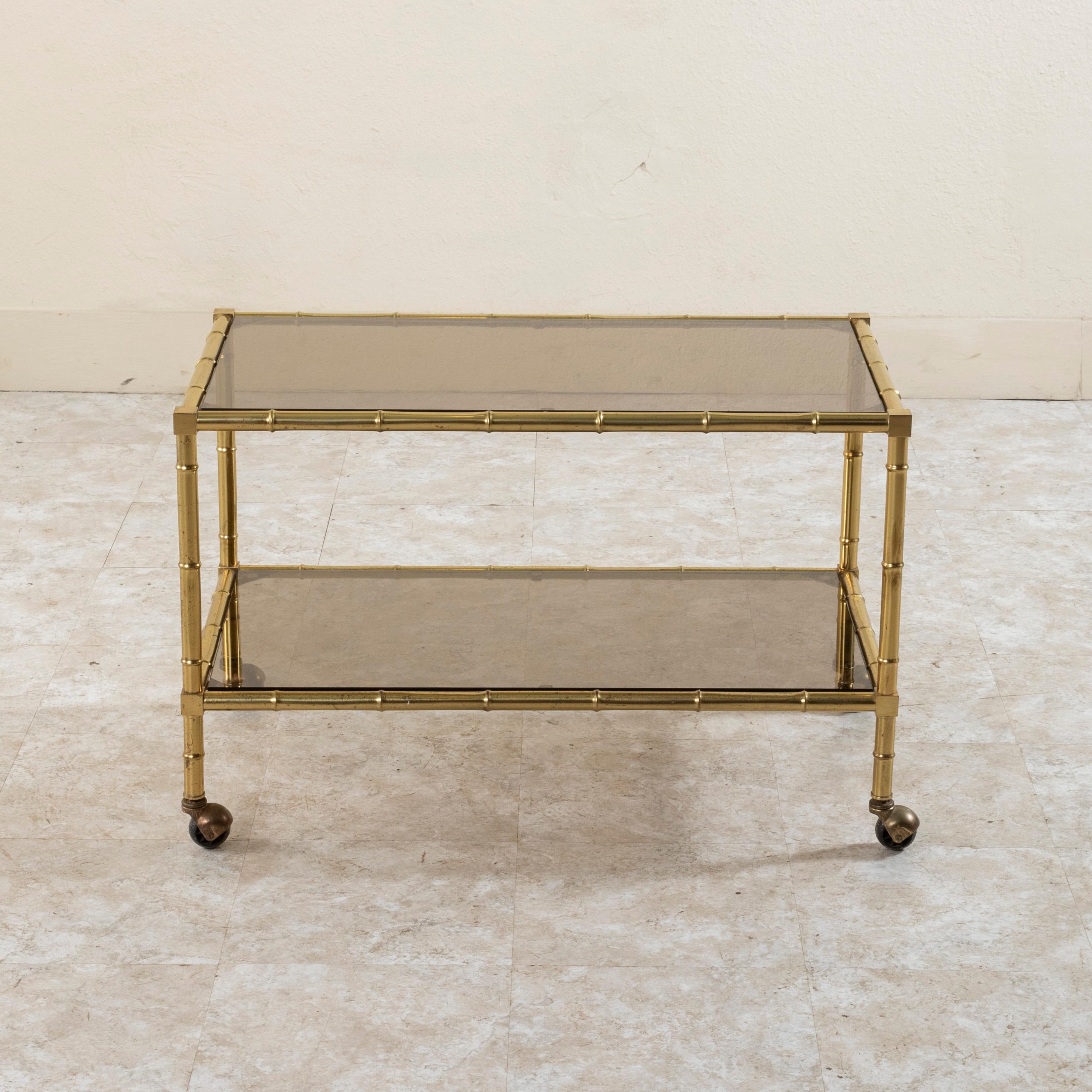 This mid twentieth century French coffee table or bar cart features a faux bamboo brass frame. Two smoked glass shelves provide ample display space. The piece rests on casters, allowing for it to be easily moved. At 20 inches in height, this piece