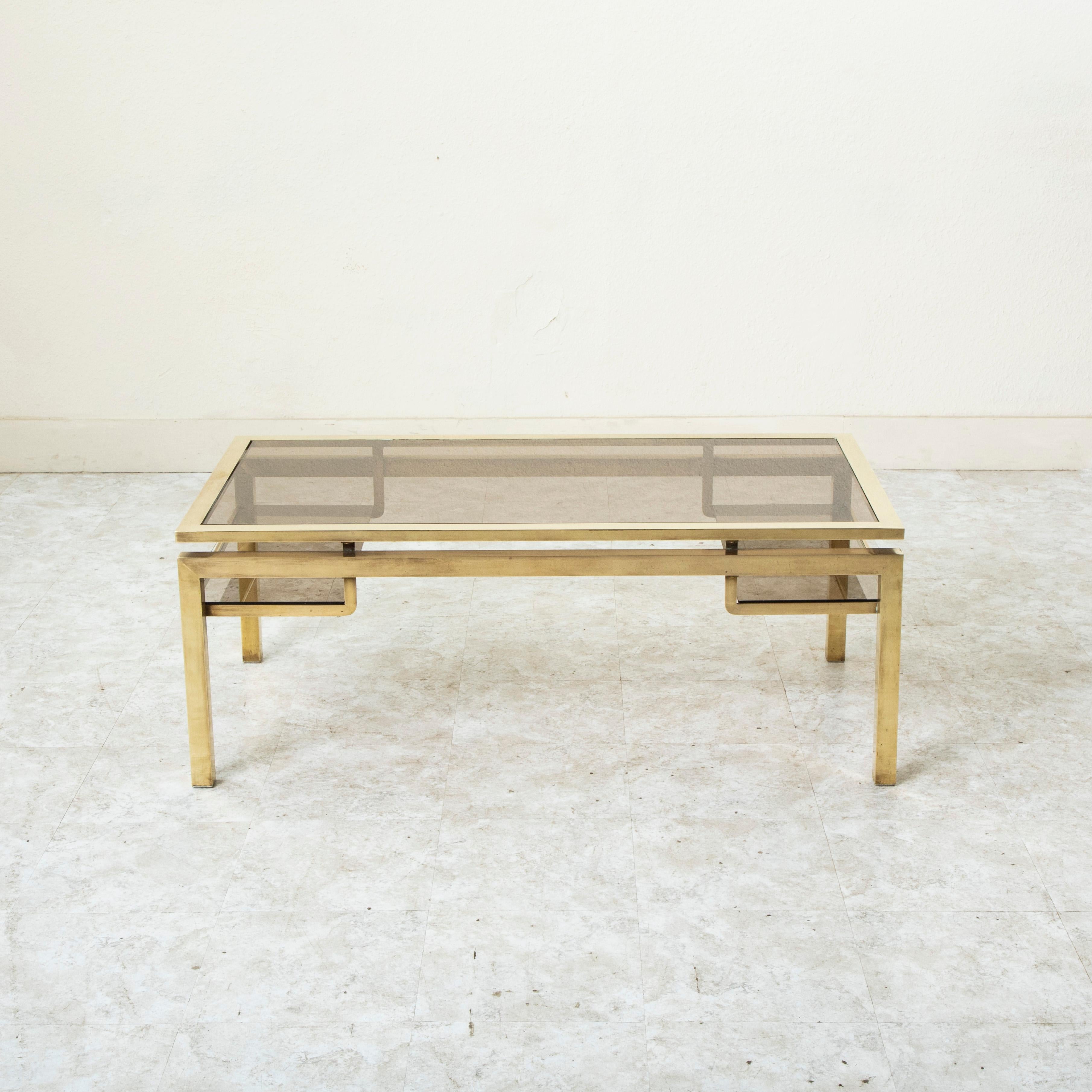 Cette table basse ou table de cocktail en laiton du milieu du siècle est attribuée au designer français Guy Lefevre. La table se compose de quatre pieds à angle droit qui sont reliés au plateau en laiton avec un insert en verre fumé. Une tablette en