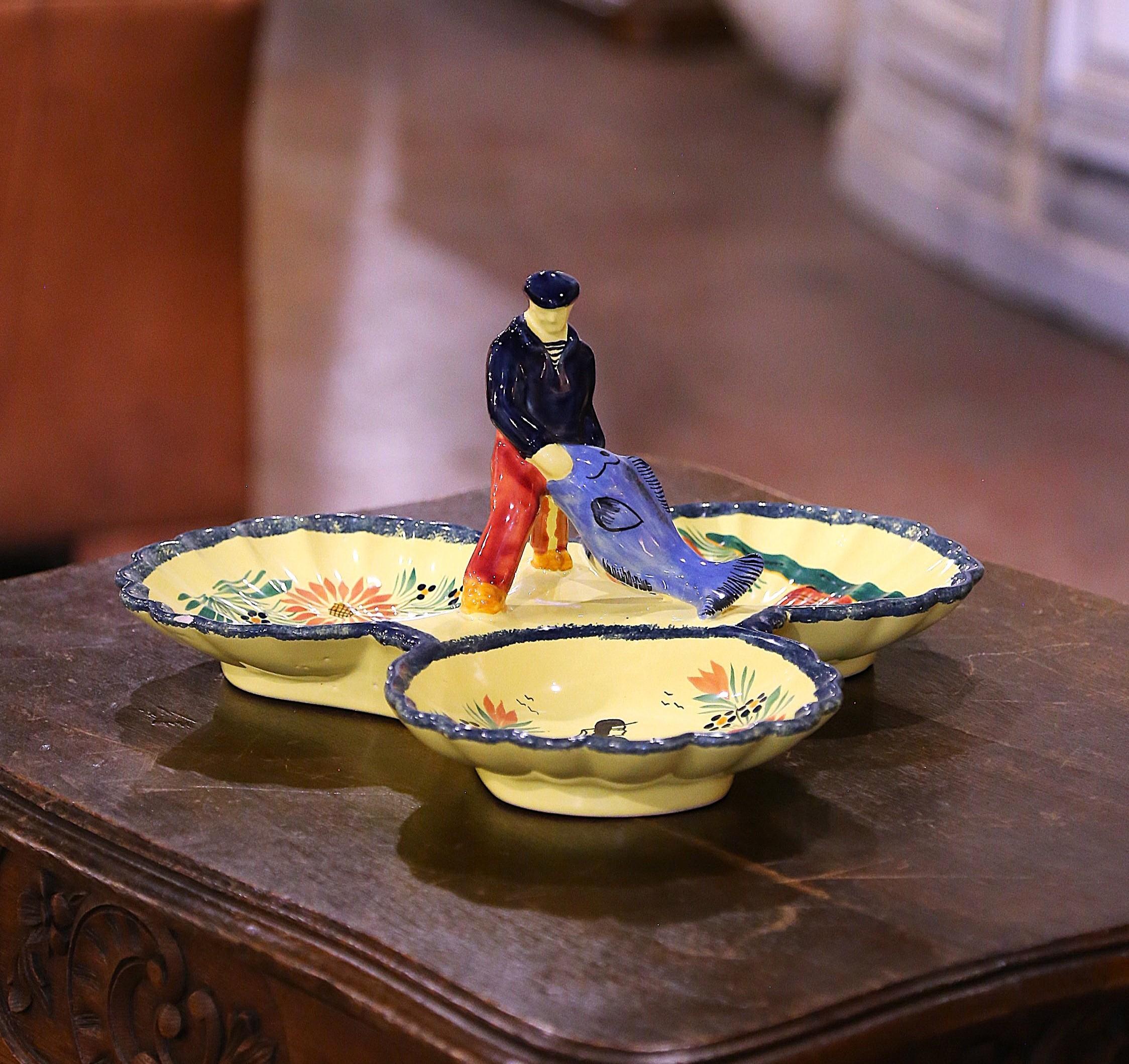 Sirve cacahuetes o patatas fritas con estilo con este colorido plato antiguo de Quimper. Esculpido en Bretaña, Francia, hacia 1950, el plato presenta una figura central esculpida de un pescador con el atuendo tradicional bretón -abrigo azul marino,