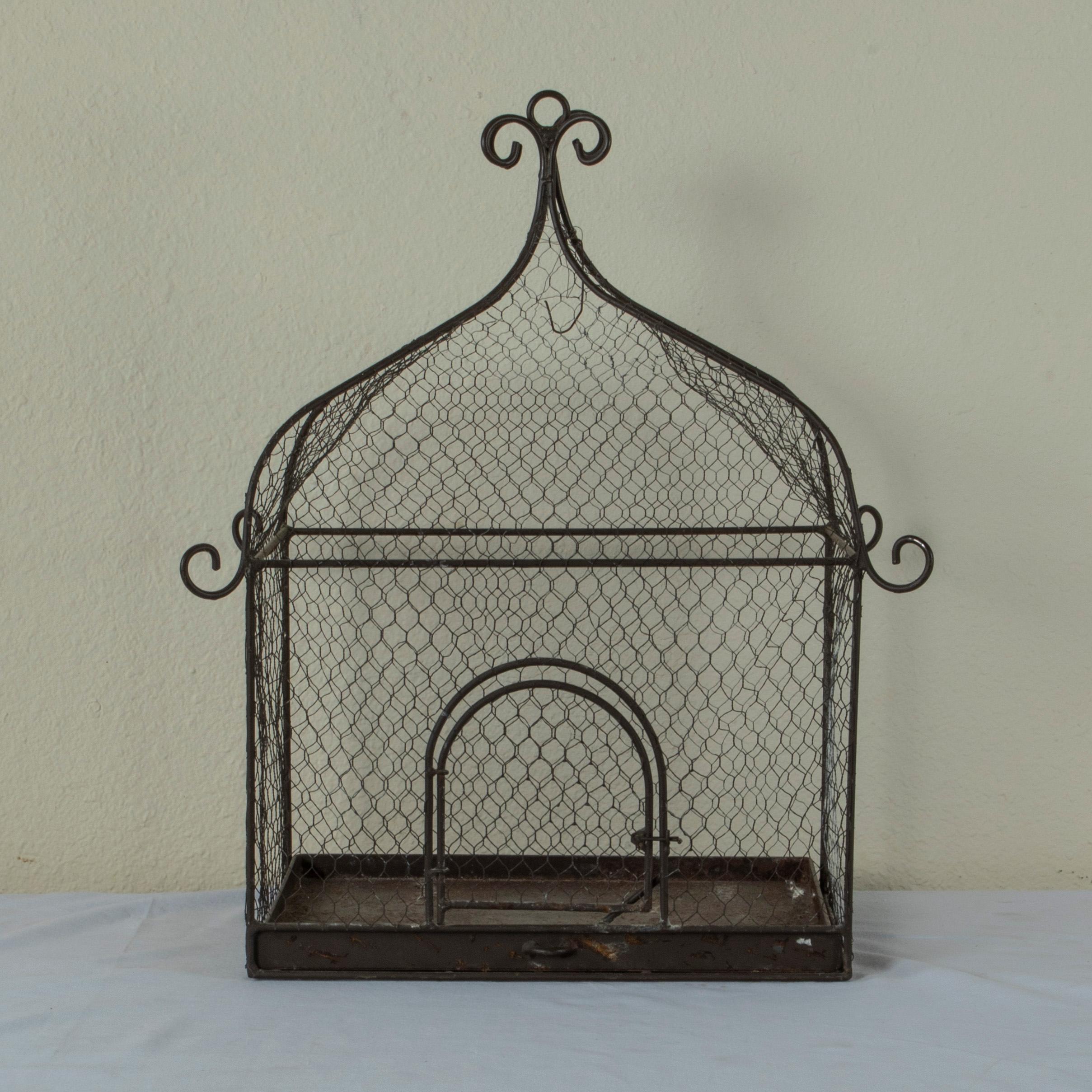 This mid-century French iron birdcage fitted with aviary wire features a pullout tray in the bottom and scrolling details at the corners and on the crown. A hinged door allows access to the interior. This birdcage may either be placed on a surface