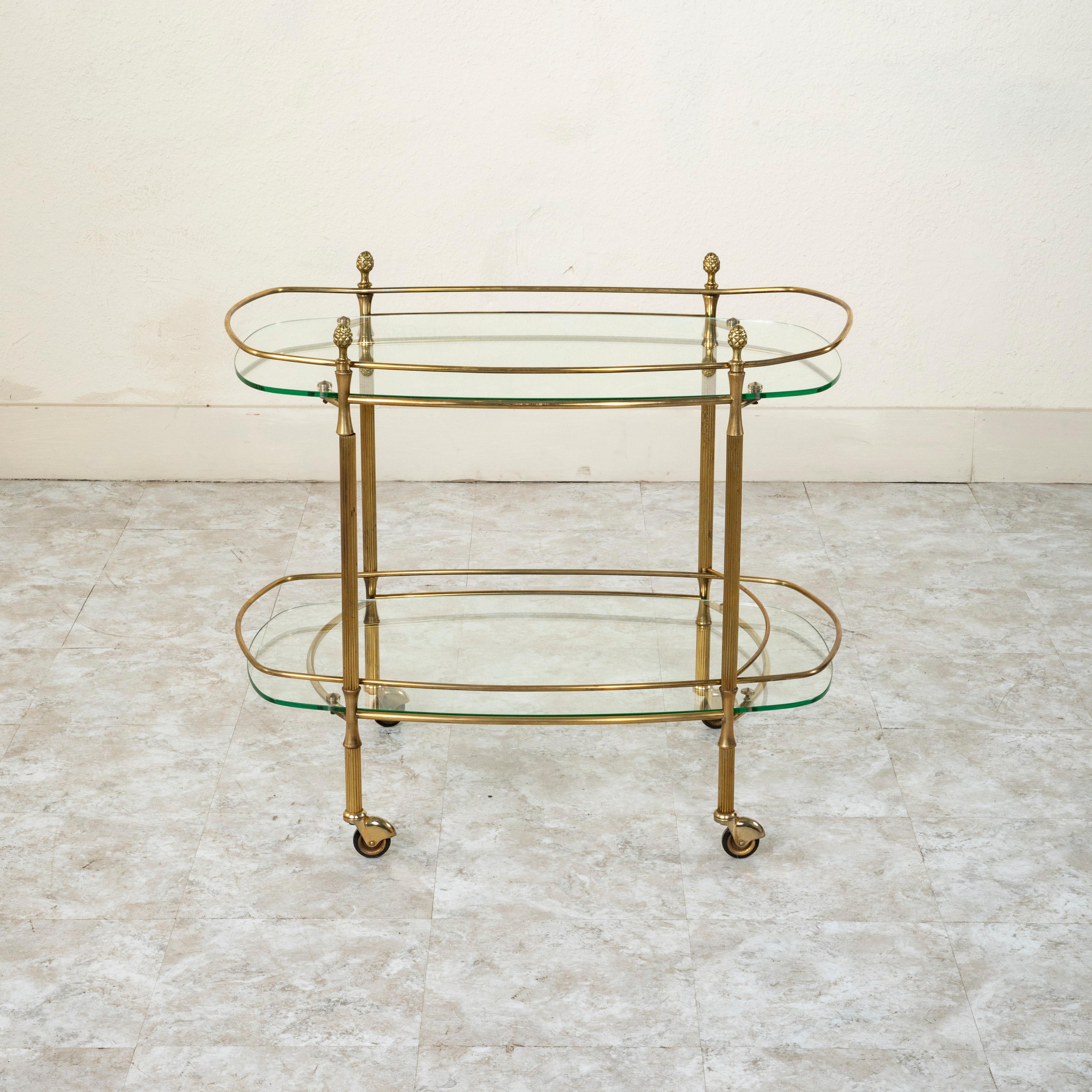 This mid-century French Louis XVI style brass bar cart features fluted columns capped with classic pinecone finials. The piece is fitted with two glass shelves each surrounded by a brass rail. The lower shelf is fitted with an additional bar that