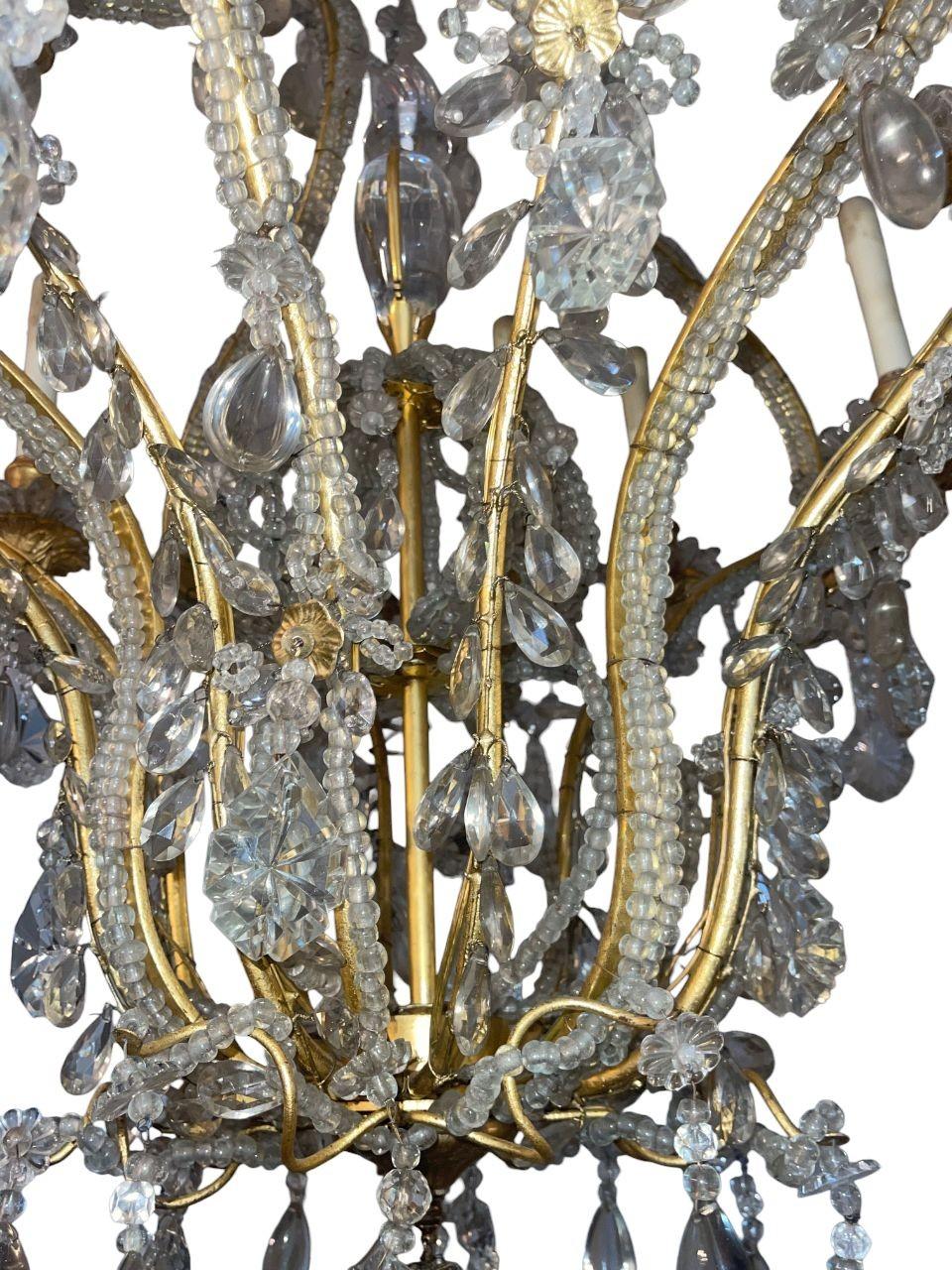Un lampadario monumentale della Maison Baguès, alto 62 pollici e con un diametro di 30 pollici, riccamente adornato con gocce di cristallo tagliate a mano, rosette e ghirlande di perline. I bracci dorati a spirale sono intricati e avvolti da fili di