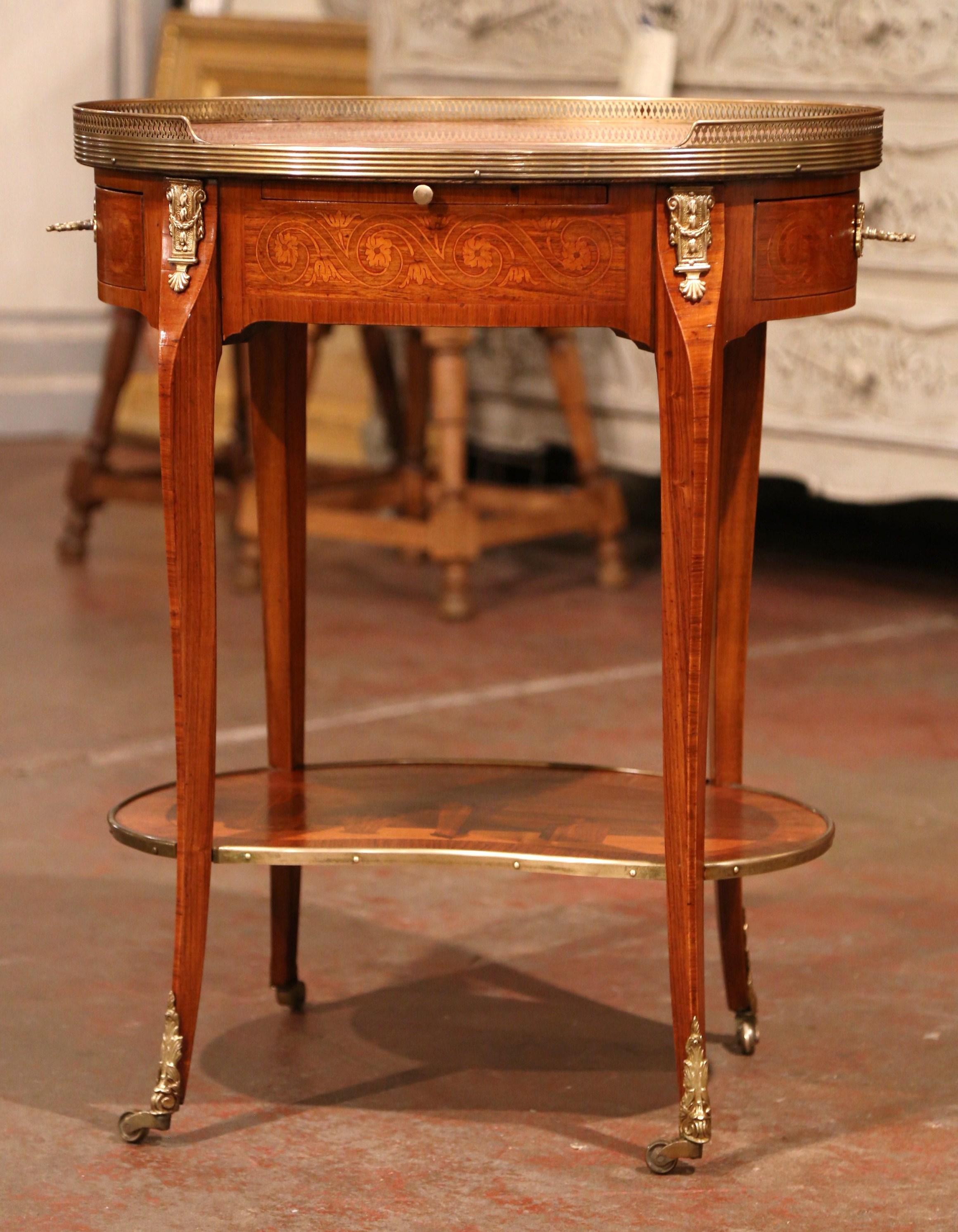 Crafted in France circa 1940 and made of cherry, the elegant table stands on four cabriole legs embellished with brass mounts over the feet and wheels at the bottom. Oval in shape, the table features detailed inlay and marquetry floral decor around