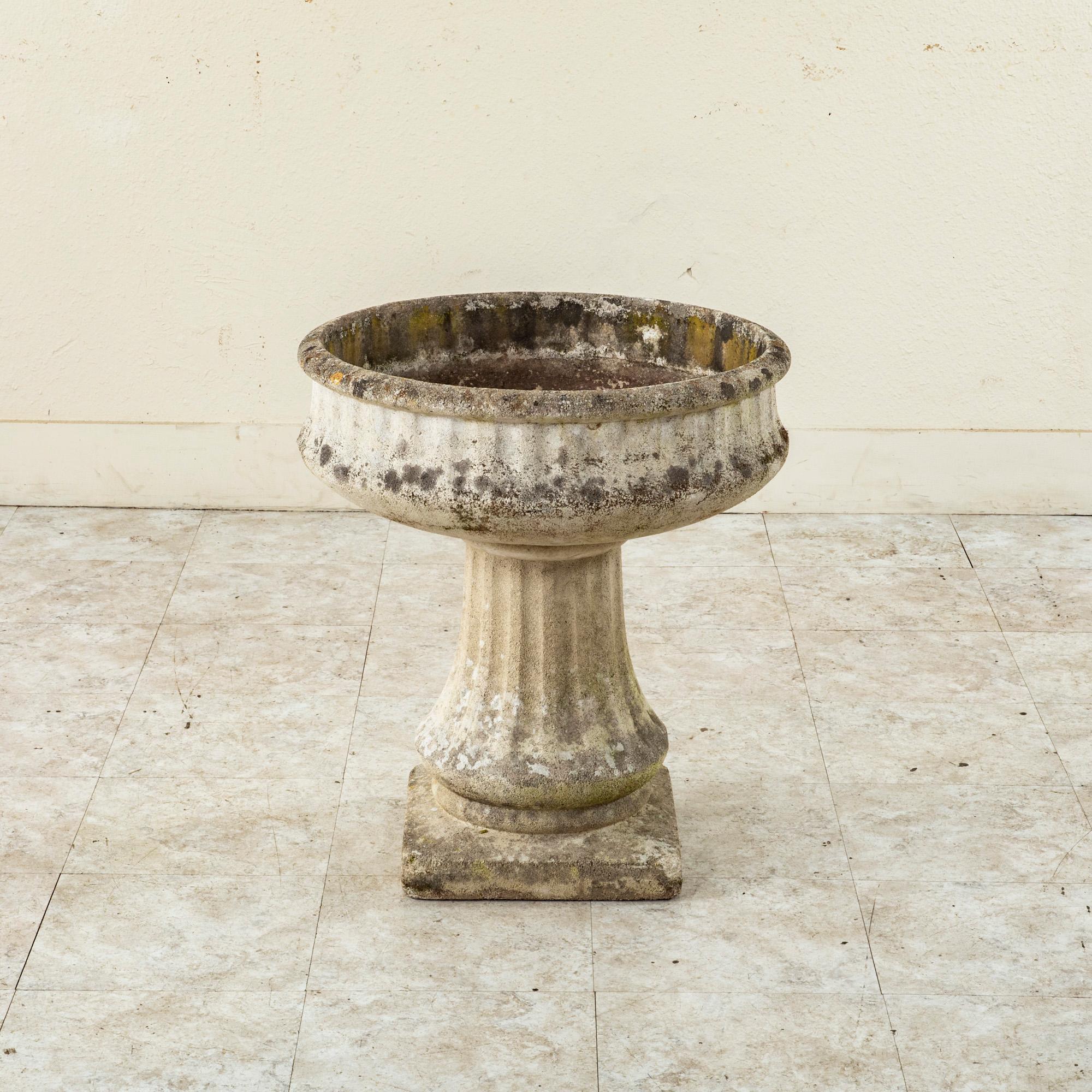 This mid twentieth century French cast stone urn features a fluted basin that rests on a fluted pedestal base. The basin measures 22 inches in diameter and the pedestal rests on a 12.5 inch square base. Vestiges of moss and lichen create a contrast