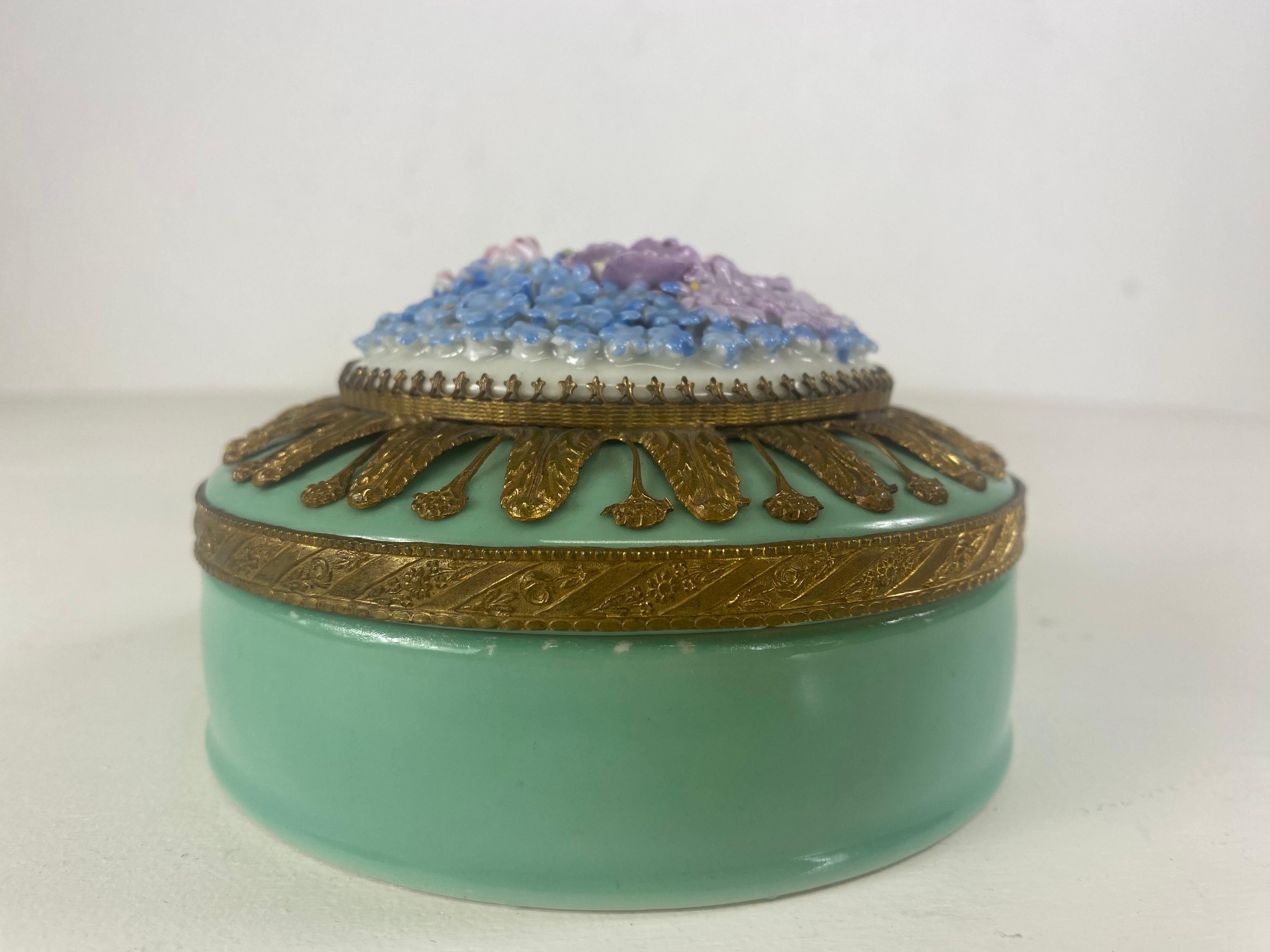 This is a mid 20th century German handmade porcelain and bronze trinket box. The trinket box has a beautiful sage green base with delicate handmade porcelain flowers on the top. The box is decorated with an ornate bronze edge at the top and around