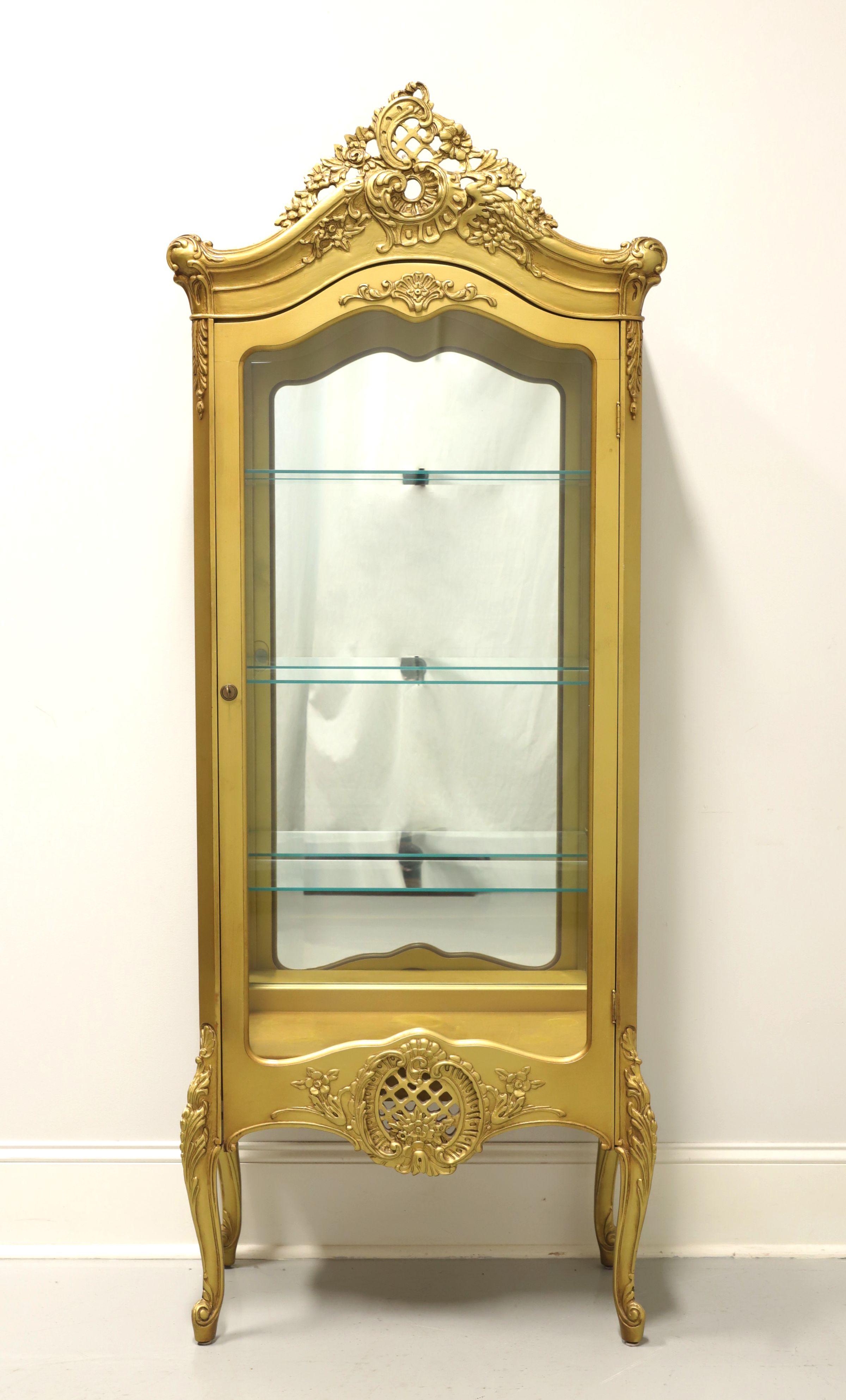 A French Country style vitrine, unbranded. Solid hardwood cabinet painted gold, brass lock hardware, elaborately carved top with center crest, glass side panels, elaborately carved apron, elevated on curved legs with carved knees and scroll feet.