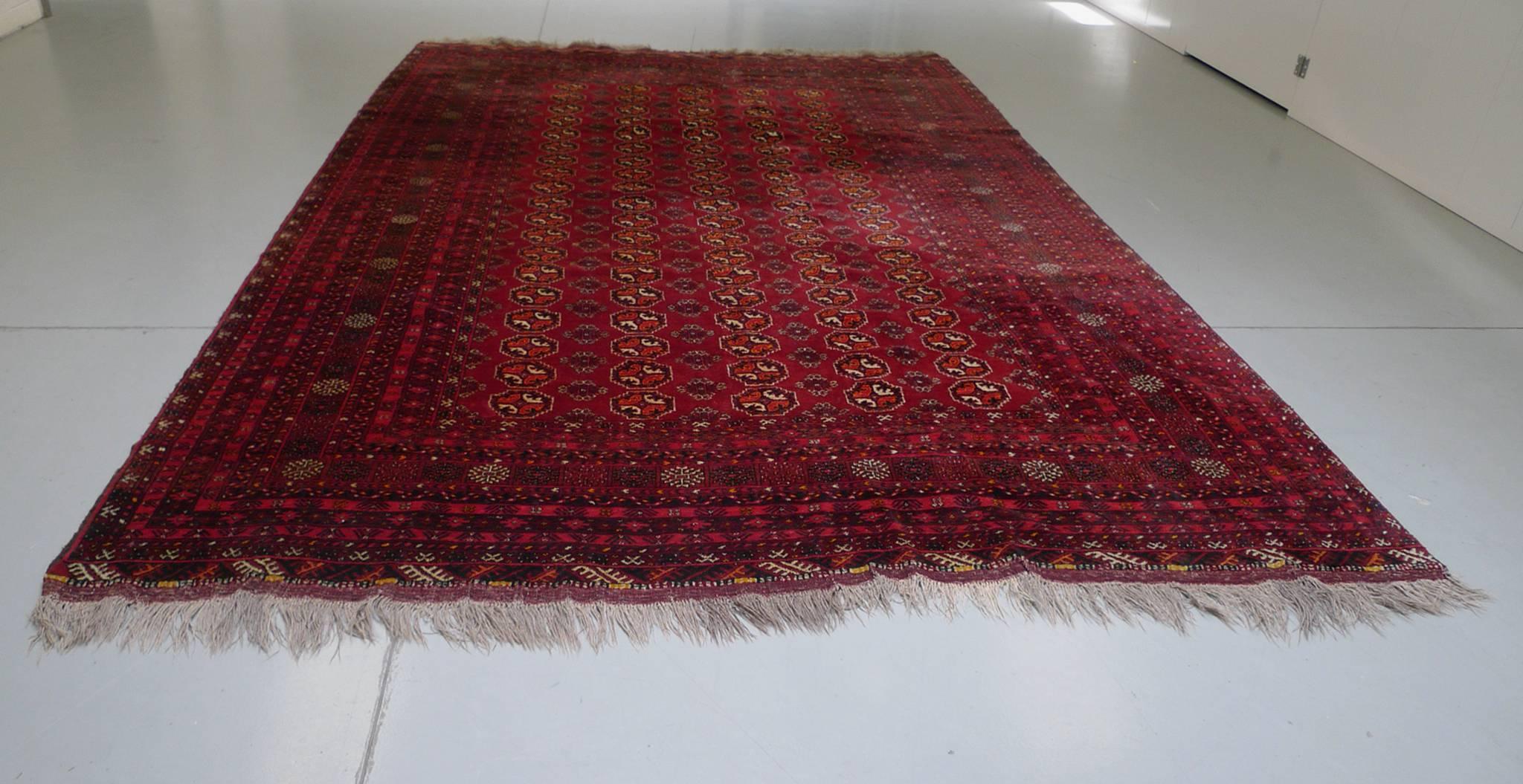 This vintage Bokhara rug was handwoven in the Mid-20th Century. Its palette is a dense mixture of deep, rich reds with ivory, blue, and orange hues hues. The design consists of five columns of traditional guls at the central rectangle, which is, in
