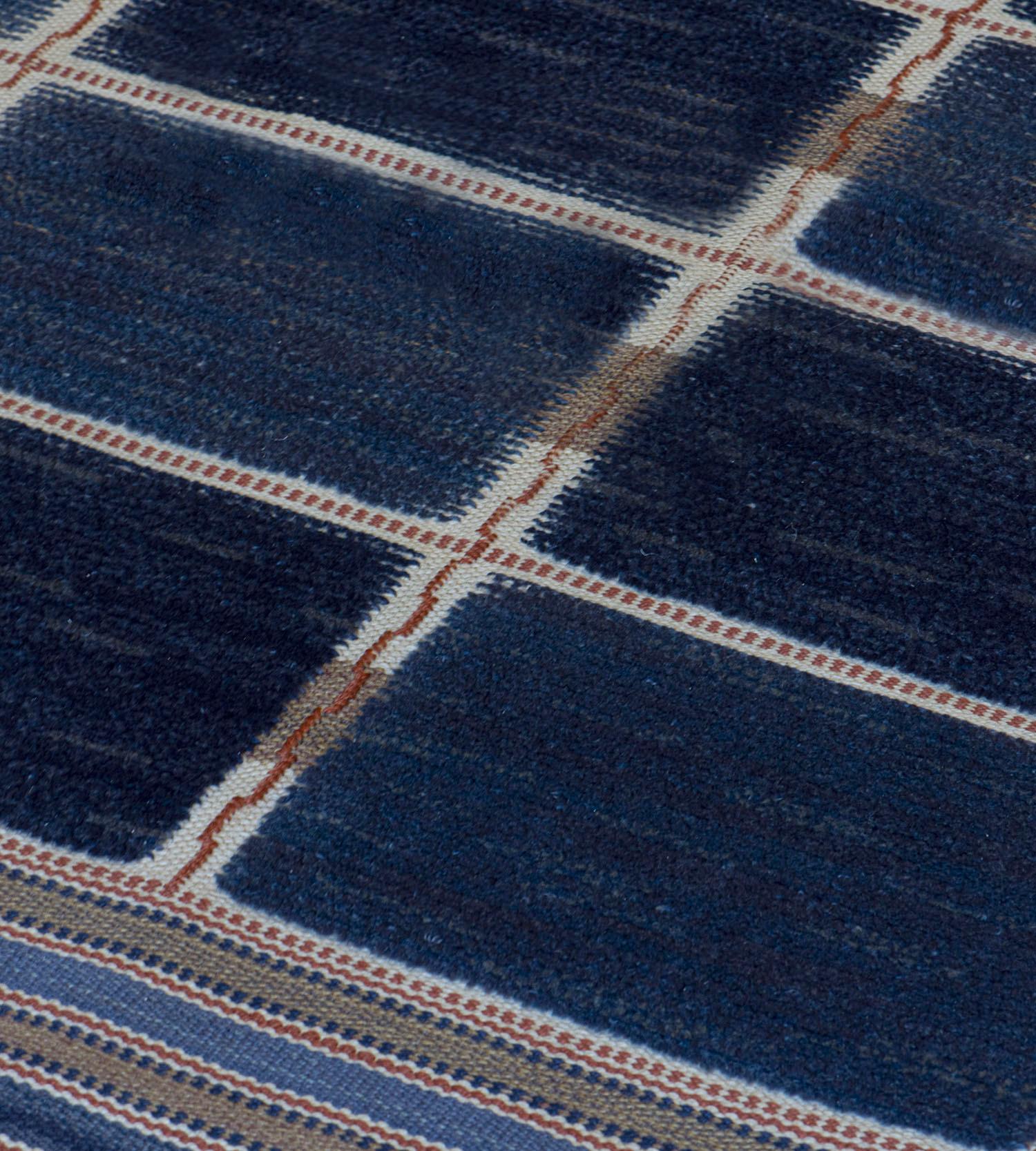 Ce tapis suédois signé présente un champ composé de huit rangées horizontales et de quatre rangées verticales de panneaux rectangulaires bleu foncé et bleu clair divisés par un cadre de rayures étroites ivoire et brun biscuit avec une ligne rouge