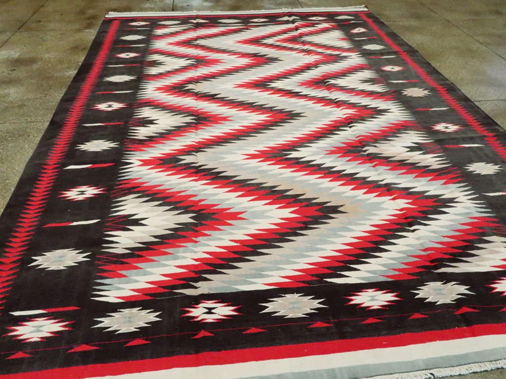 Un tappeto indiano vintage a trama piatta Dhurrie. Un audace zig-zag orizzontale in rosso, nero ed ecrù, in stile Navajo, fa muovere l'occhio su questo tappeto piatto indiano tessuto ad arazzo. Il bordo principale nero mostra gli ashik separati da
