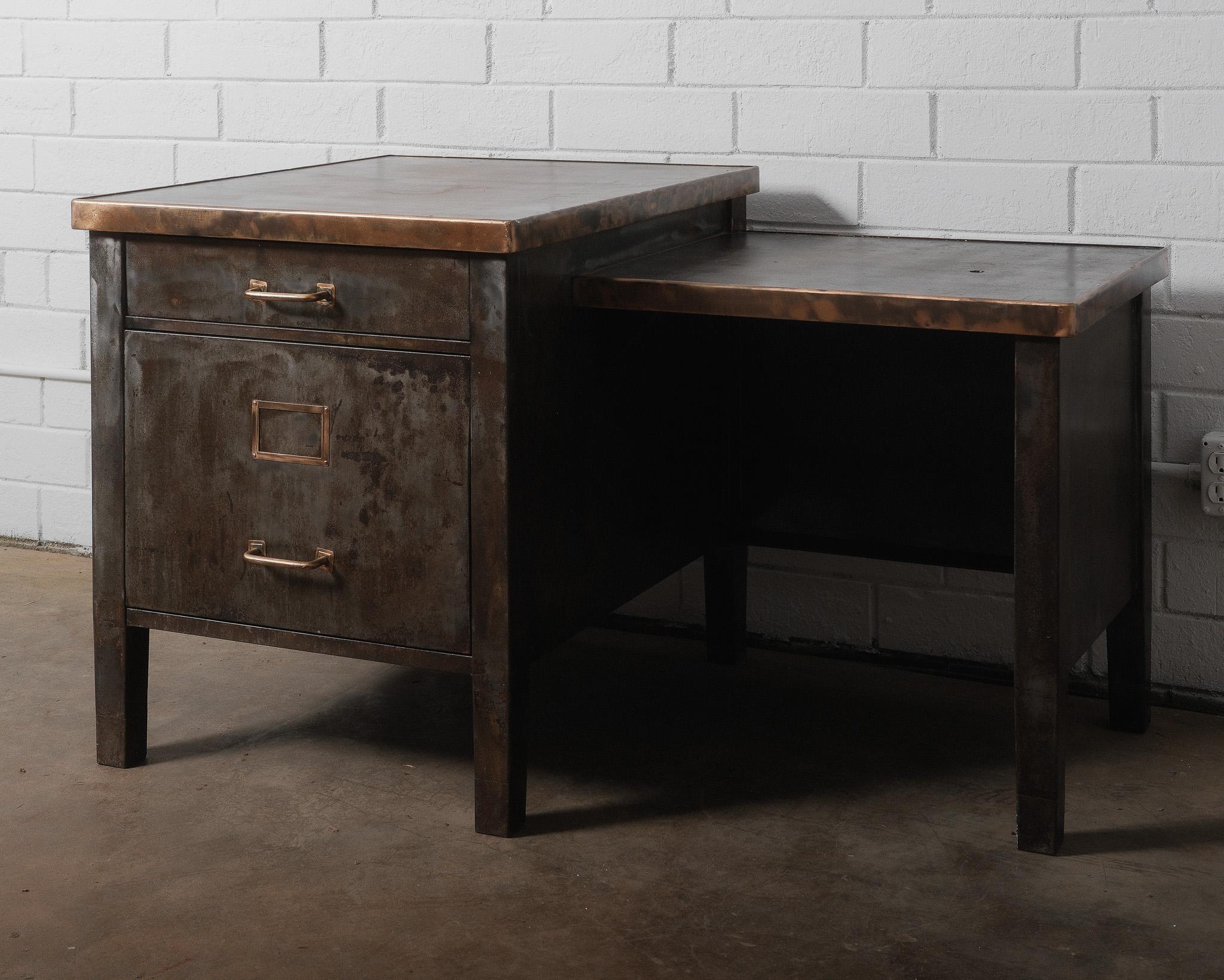 This robust industrial desk features a striking blend of aged steel construction and a warm copper-edged top, offering a distinctive look with strong visual weight. The subtle L-shaped layout includes a filing cabinet base with copper or brass-toned