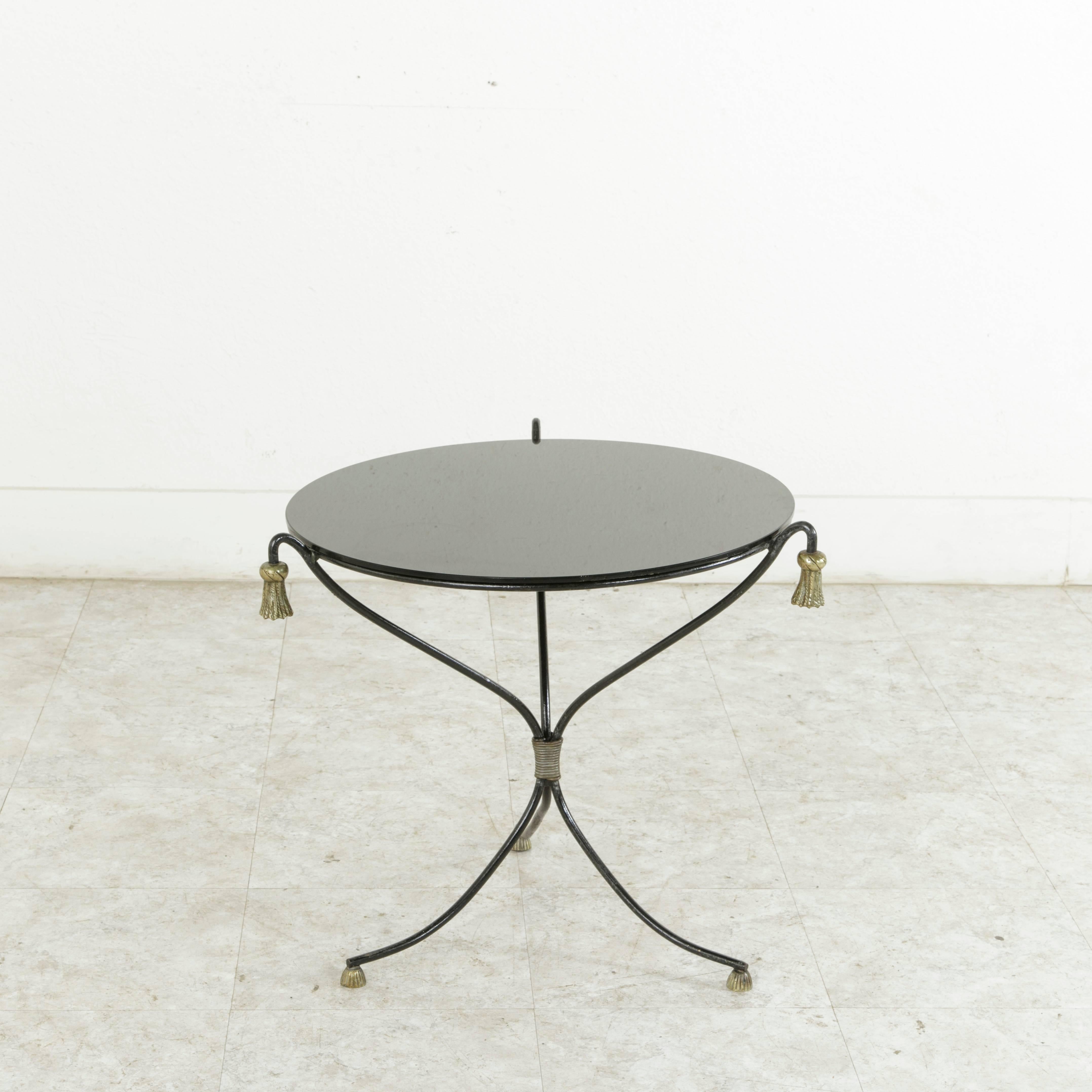 This midcentury French round iron cocktail table or side table features a pedestal base with three curved legs finished in bronze tassels both top and bottom. Its original black glass top completes the look. With a diameter of 20.5 inches, this