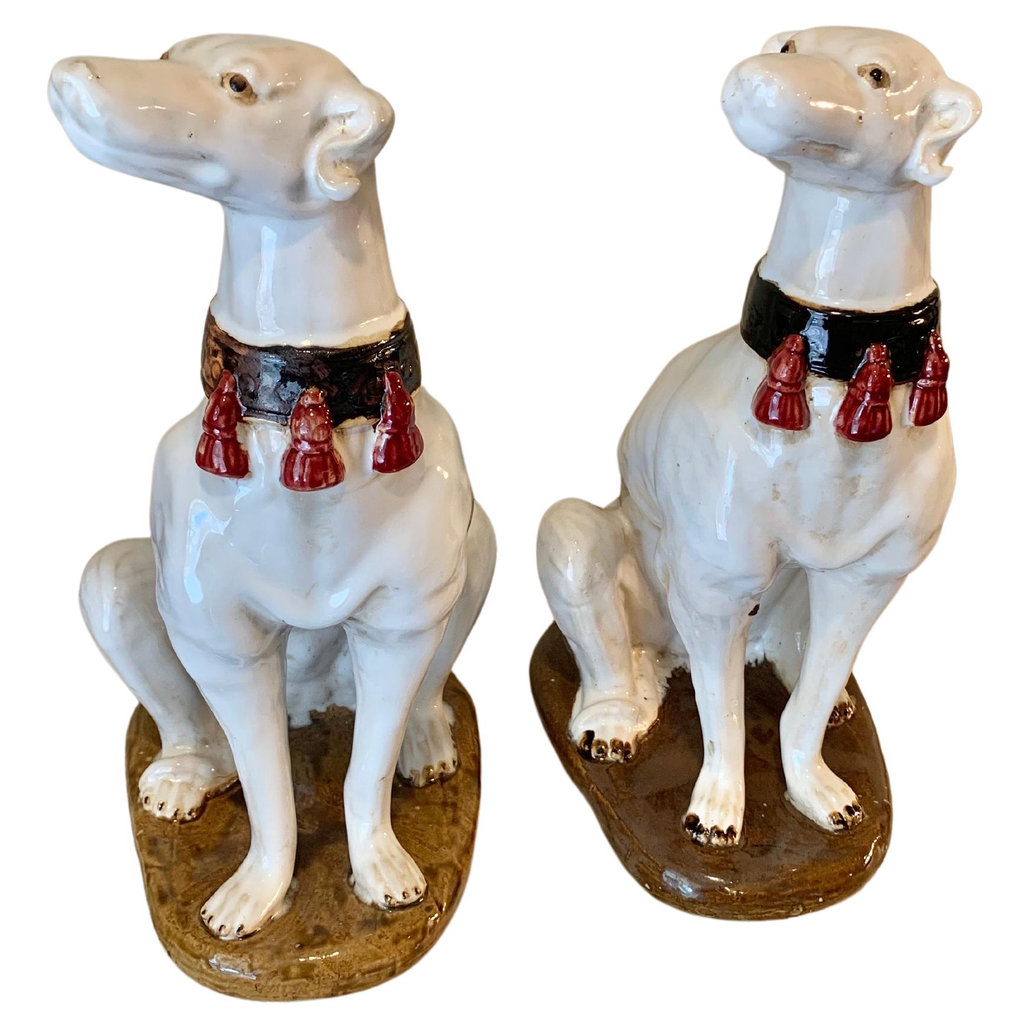 An absolutely stunning pair of Mid 20th Century Italian Ceramic Whippet Sculptures. These pottery whippets are presented with a thick black collar and red tassels against a white glazed body. They are each sitting upright on a brown base. These