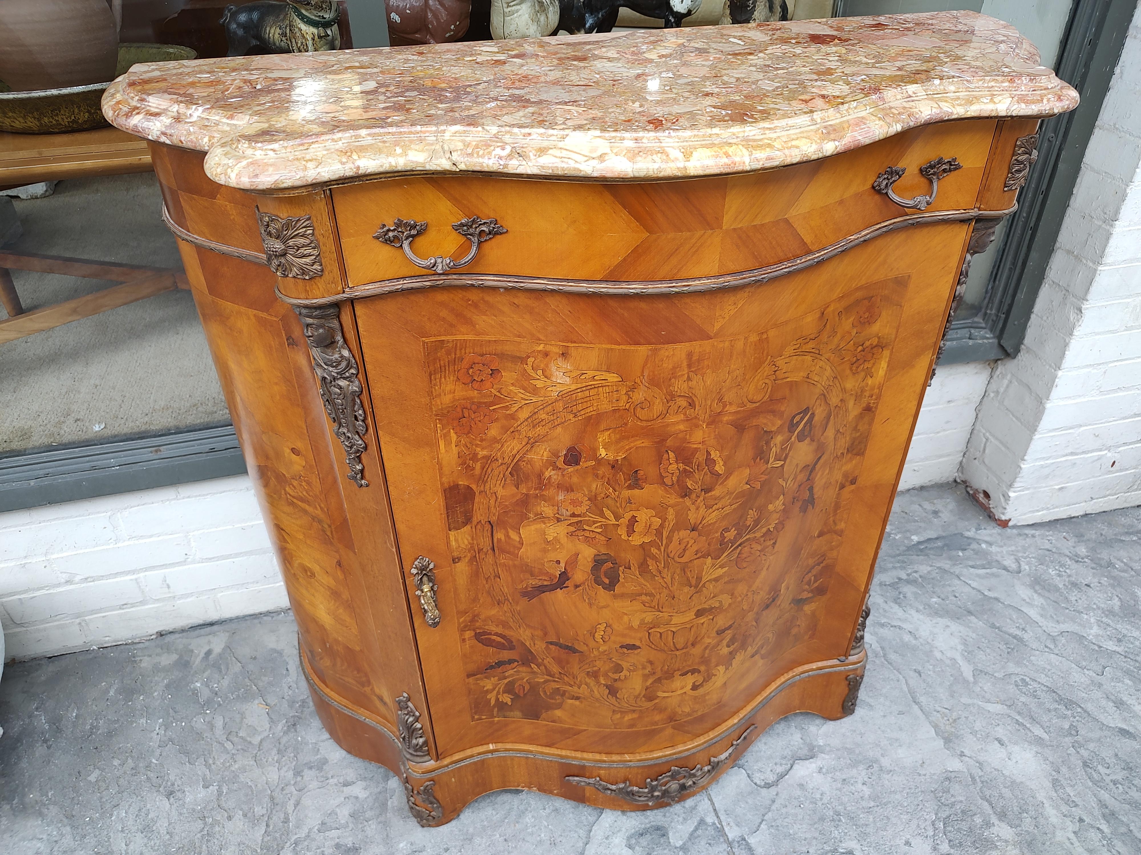 Fabulous marble top cabinet with a serpentine front and a large paneled Marquetry door. Bronze figural mounts, lock with working key. Marble was cracked and repaired, see pics. Interior has a shelf for added storage. In excellent vintage condition