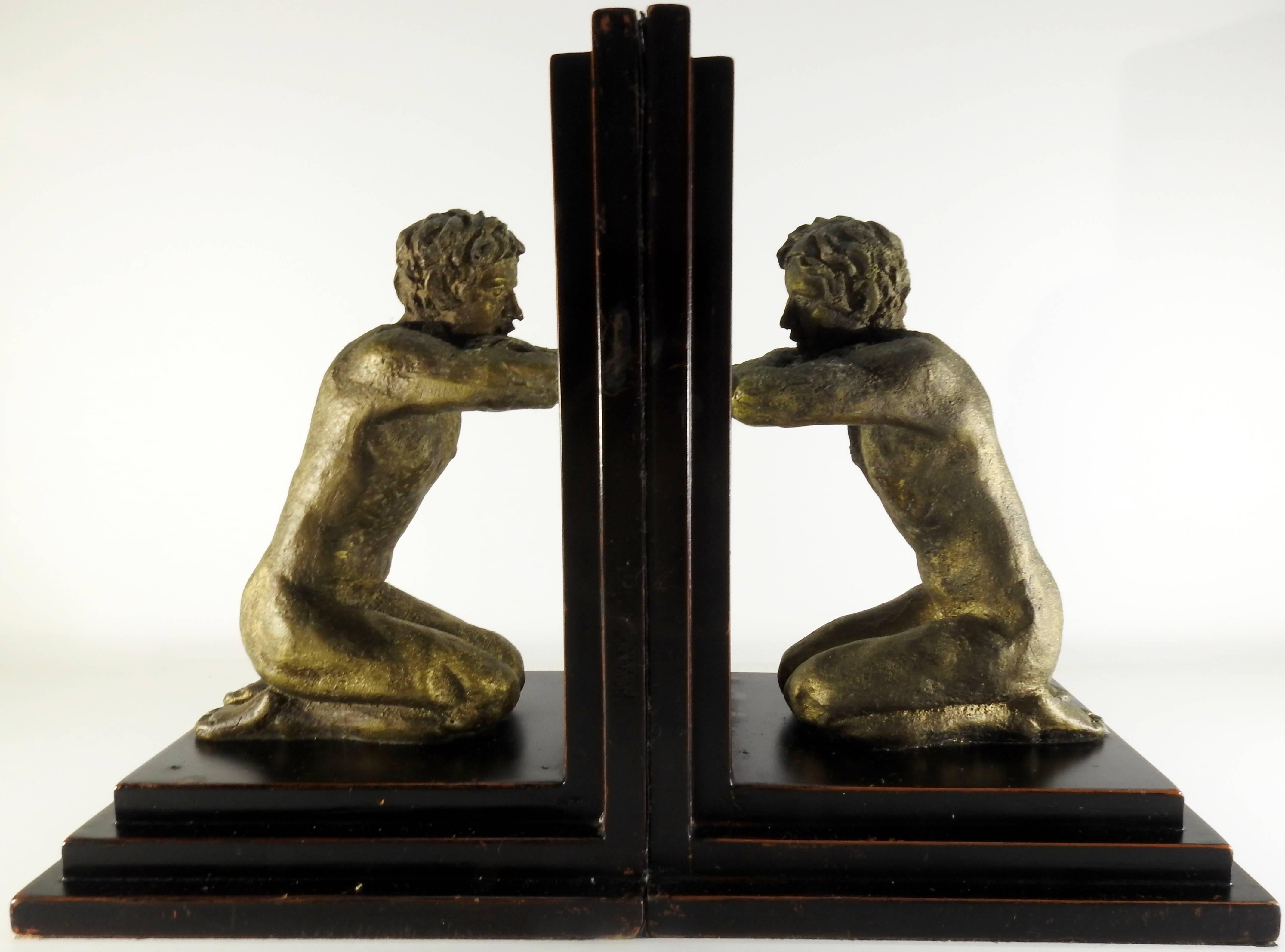 A pair of kneeling men are gazing in their respective mirrors in this pair of midcentury bookends. The nude men have rustic details and are finished in an antique gold color. They are mounted on stair step wooden bases. The bottoms and sides are