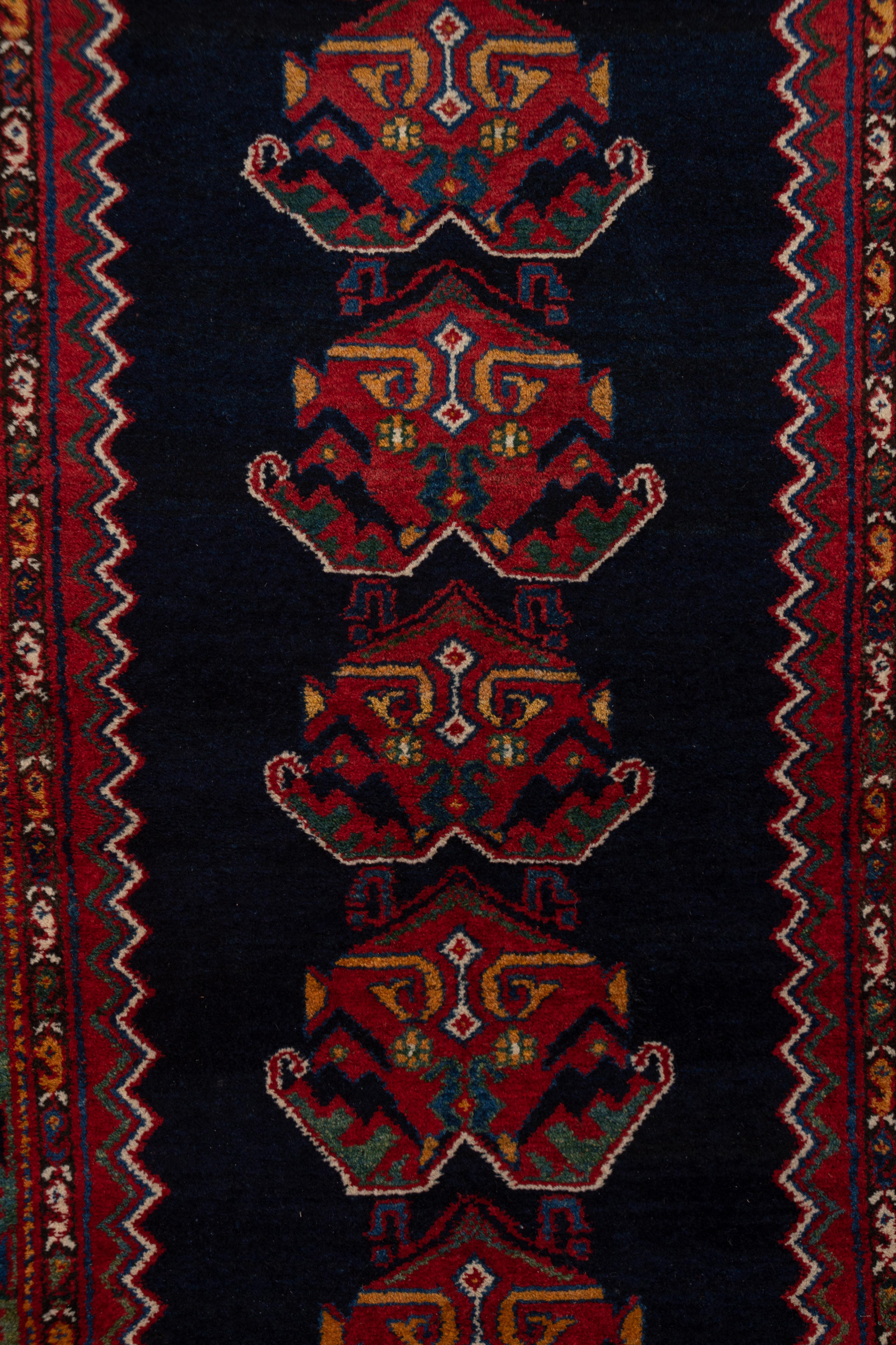 This west Persian runner displays a zig-zag edged navy otherwise plain sub field with 19 leafy palmettes, on a red ground. The green main border displays small ashik medallions between botteh chain guards.