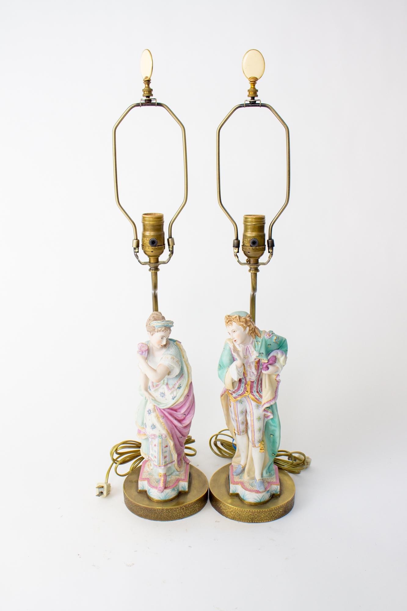 Mid 20th century masquerade bisque figurine lamps. A pair featuring a man and a woman in pastel tones, the woman wearing a pink cape and the man wearing a turquoise cape, both holding masks as they reveal their faces. Bisque or kalk porcelain,