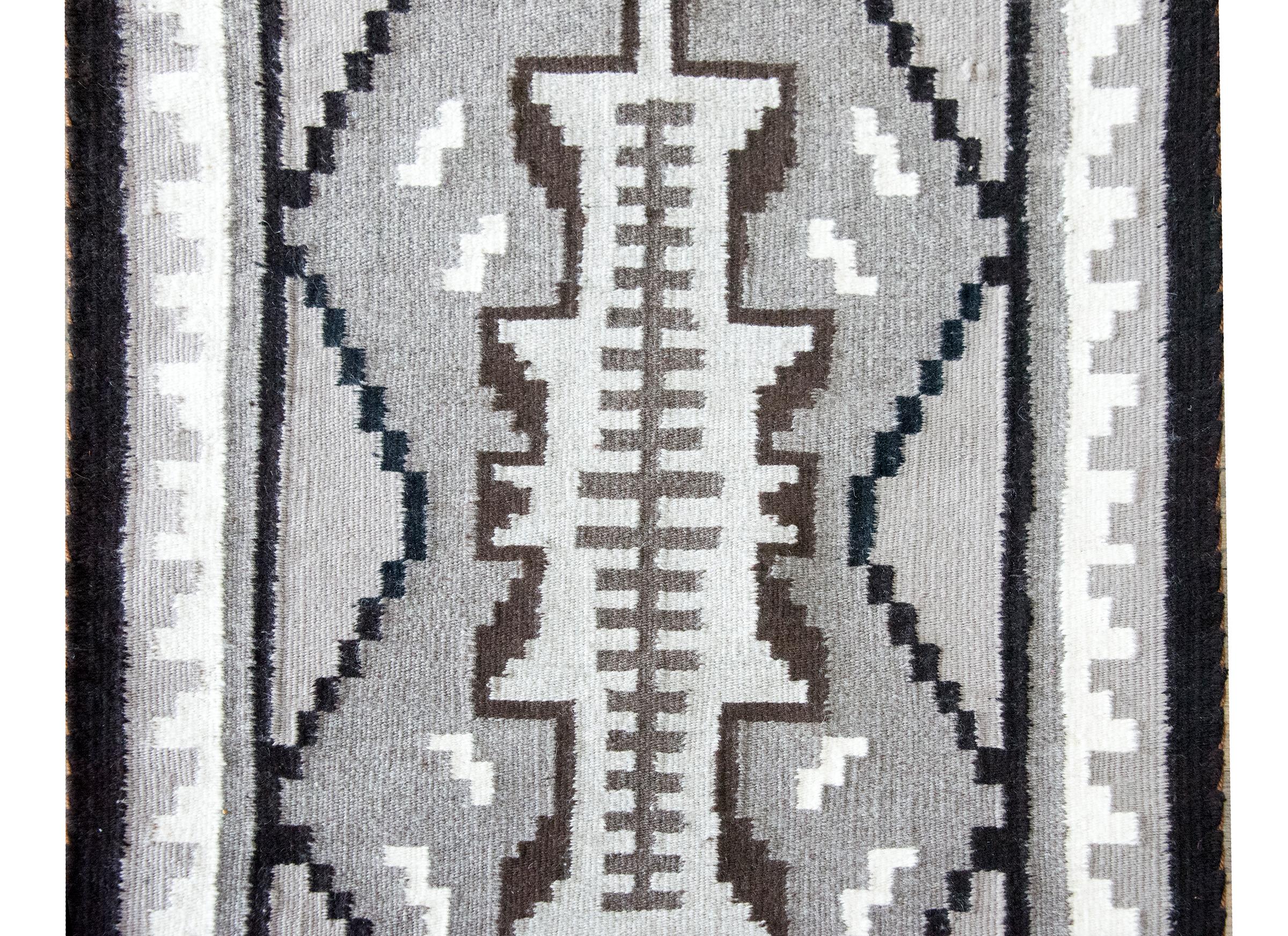 Questo tappeto Navajo della metà del XX secolo si distingue per la sua sorprendente tavolozza monocromatica e per l'insolito medaglione centrale. Allungato e stretto, il medaglione presenta un motivo a strisce semplice ma ritmico che attira lo
