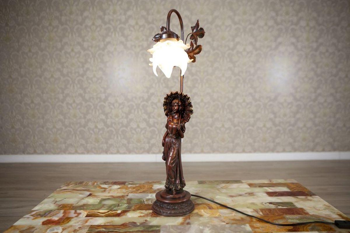 20th-Century Night Light Featuring Woman Figurine Made of Zamak Alloy

A night light made of patinated zamak alloy. The base features a figurine of a woman with an umbrella, while the upper part is adorned with floral motifs. The item dates to the