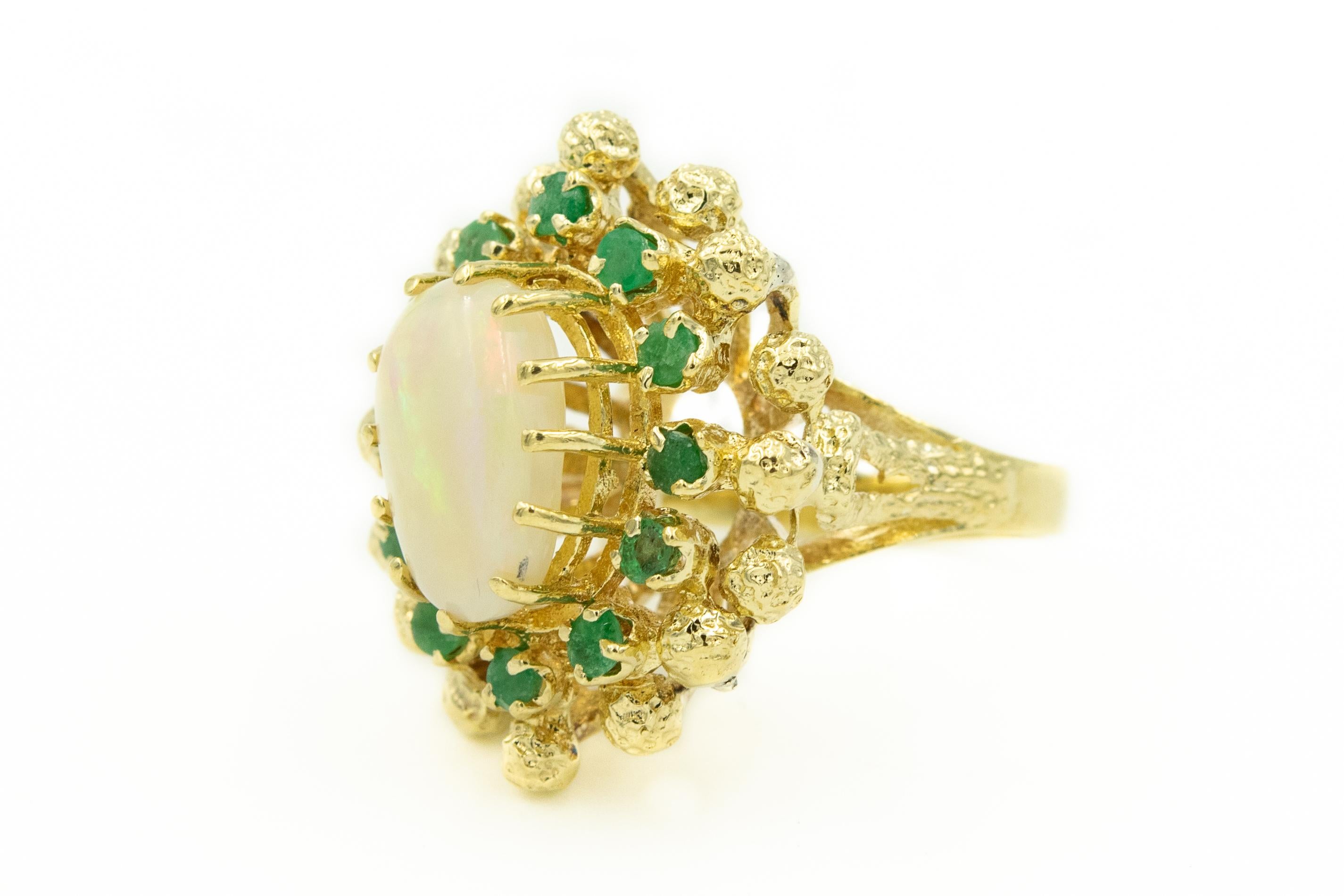 Organic ring from the 1960 - 1970s era, this cluster style ring features an oval opal with a beautiful red, blue, green and yellow flat of color set among 12 prong set emerald with an outer rim of textured balls.  The ring is 14k yellow gold.  

US