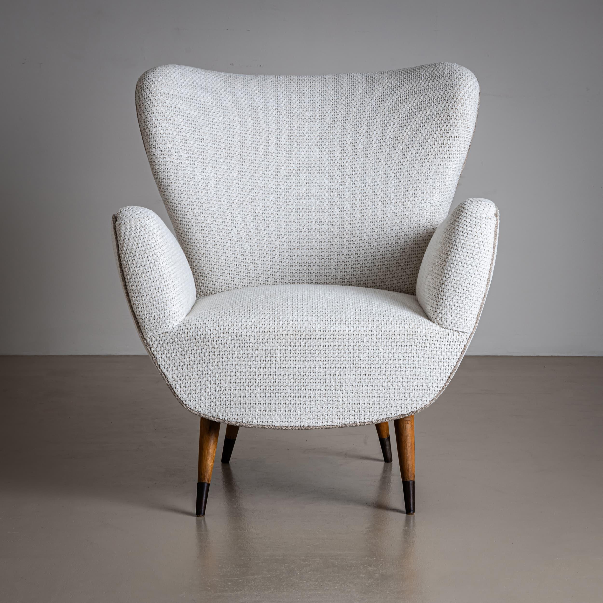 Elegante coppia di poltrone imbottite con schienali trapezoidali e sedute a U con braccioli alti e dolcemente curvati. Le sedie poggiano su gambe affusolate in legno con punte annerite, che aggiungono un tocco raffinato al design. Sono stati