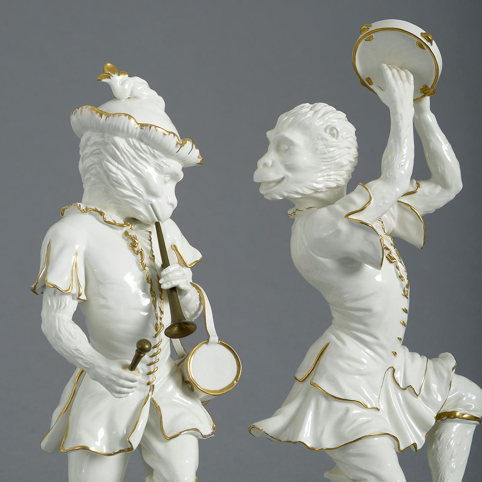 A pair of mid 20th century Italian Singerie porcelain monkey musicians one playing a tambourine, the other a drum and trumpet, shown in 18th century costume in white heightened with gilding.