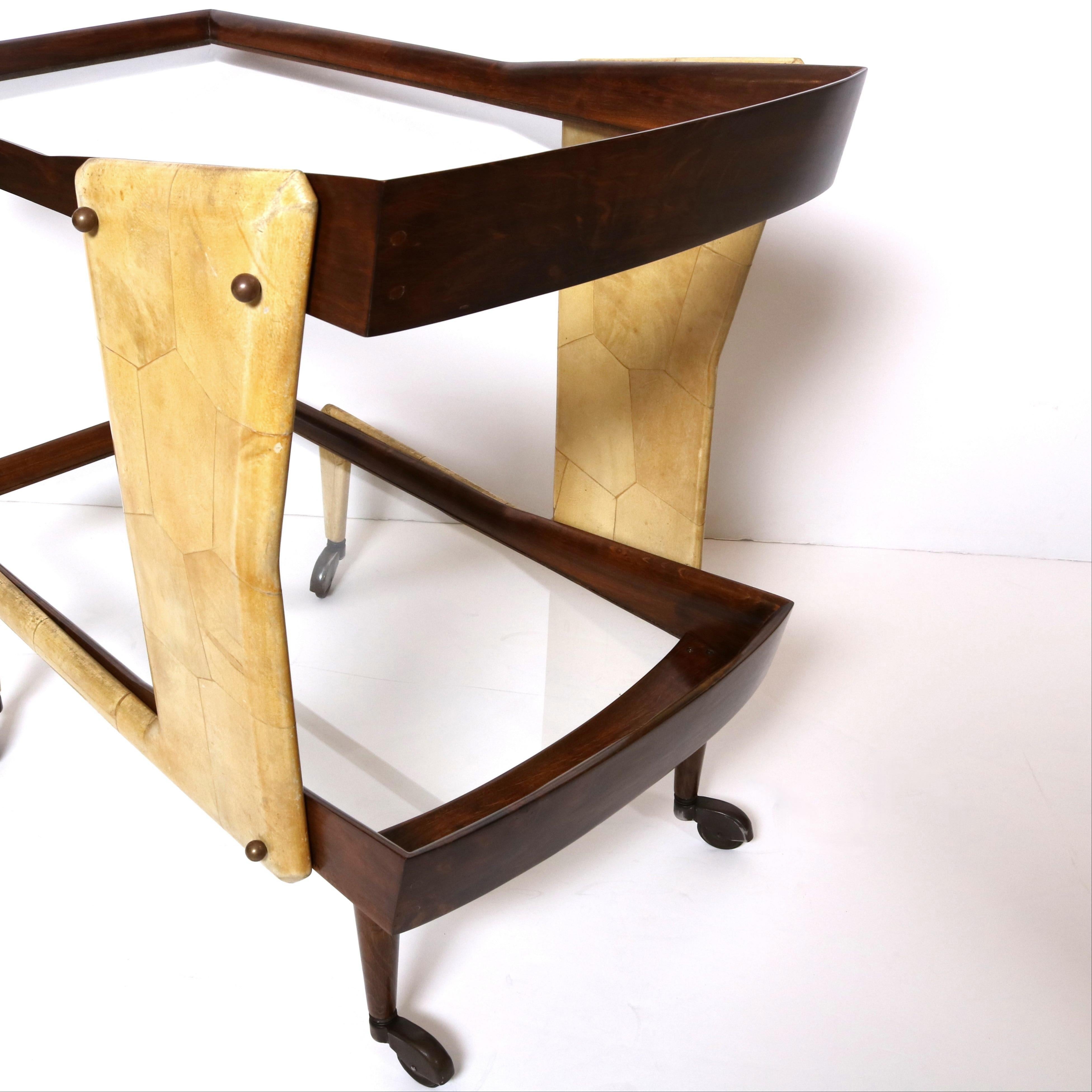 Carrello da bar a sbalzo in pergamena, legno e vetro.  Questo elegante carrello da bar presenta due ripiani in vetro incorniciati da una cornice in legno scolpito e tinto scuro.  Il legno è contrastato da gambe d'epoca rivestite di pergamena che si