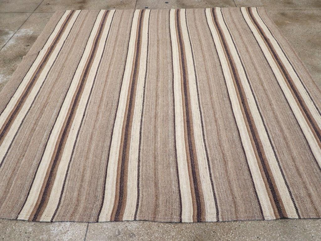 Esta alfombra cuadrada persa Flatweave Kilim de mediados del siglo XX, que mide 3,8 m x 3,8 m, presenta un diseño a rayas atemporal que desprende calidez y sencillez. La composición está definida por bandas verticales alternas en tonos terrosos de
