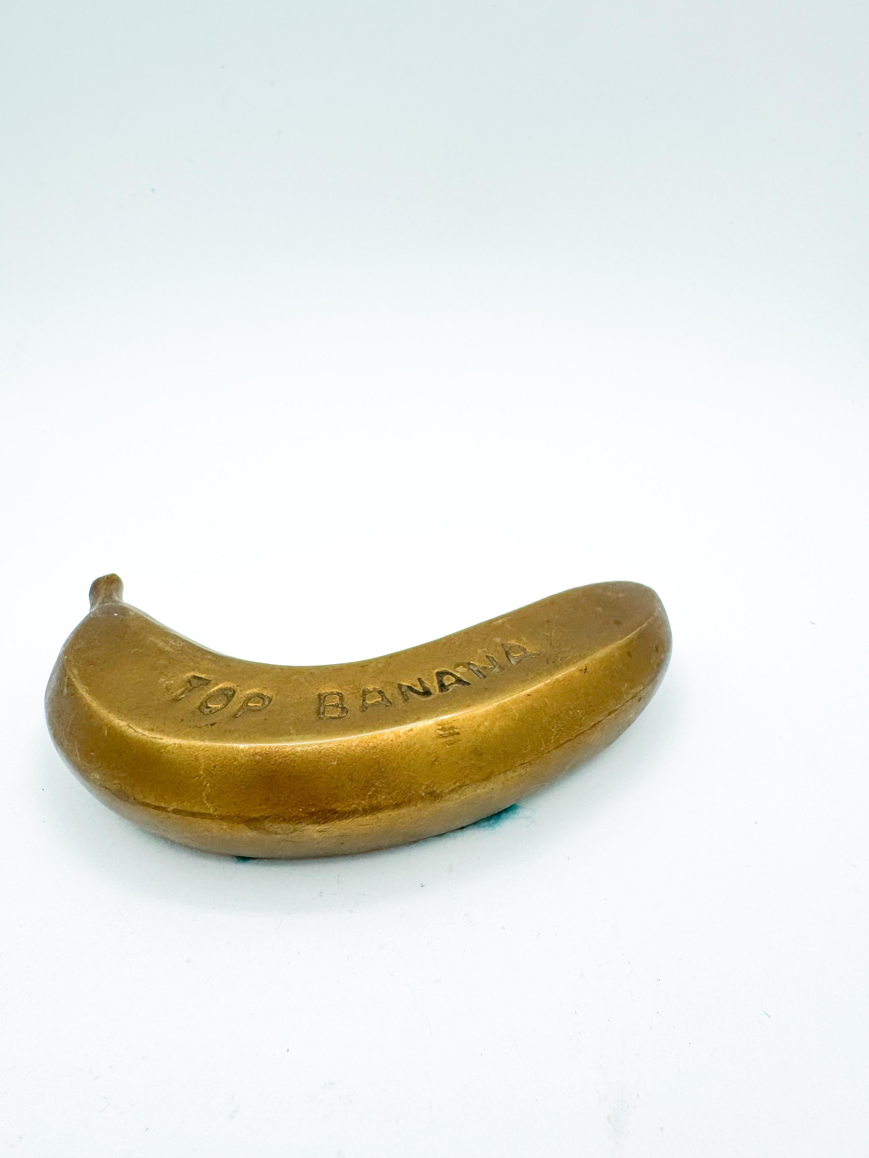 A fun and funky piece of desk or coffee table decor, this Mid 20th Century solid brass “Top Banana” paperweight is a quirky nod to pop art and playful design. Engraved with the phrase “TOP BANANA” across its curved body, it celebrates leadership and