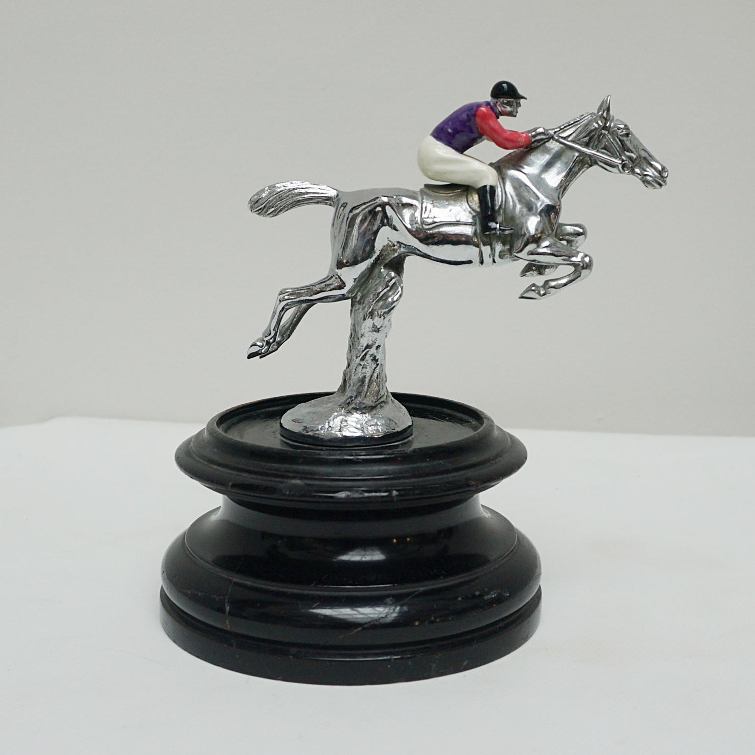 A midcentury racing horse and jockey, jumping over a hurdle. chromed metal with enamel painted Queens colours. Set over circular wooden base.