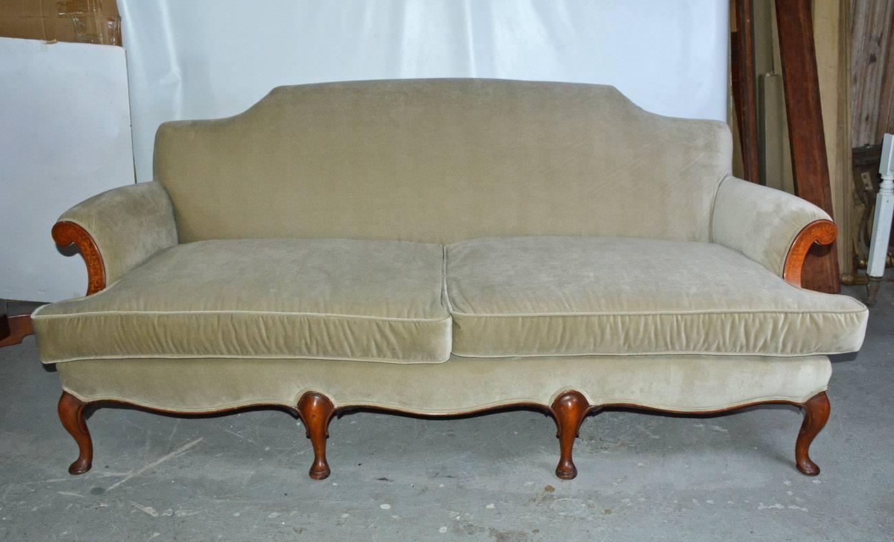 The mid-20th century sofa has wood cabriole legs with padded feet set within undulating front and side edges. The upholstered arms are fronted by inlaid curved wood panels. Seats four. The cotton velvet fabric is in a light shade of grey-green.