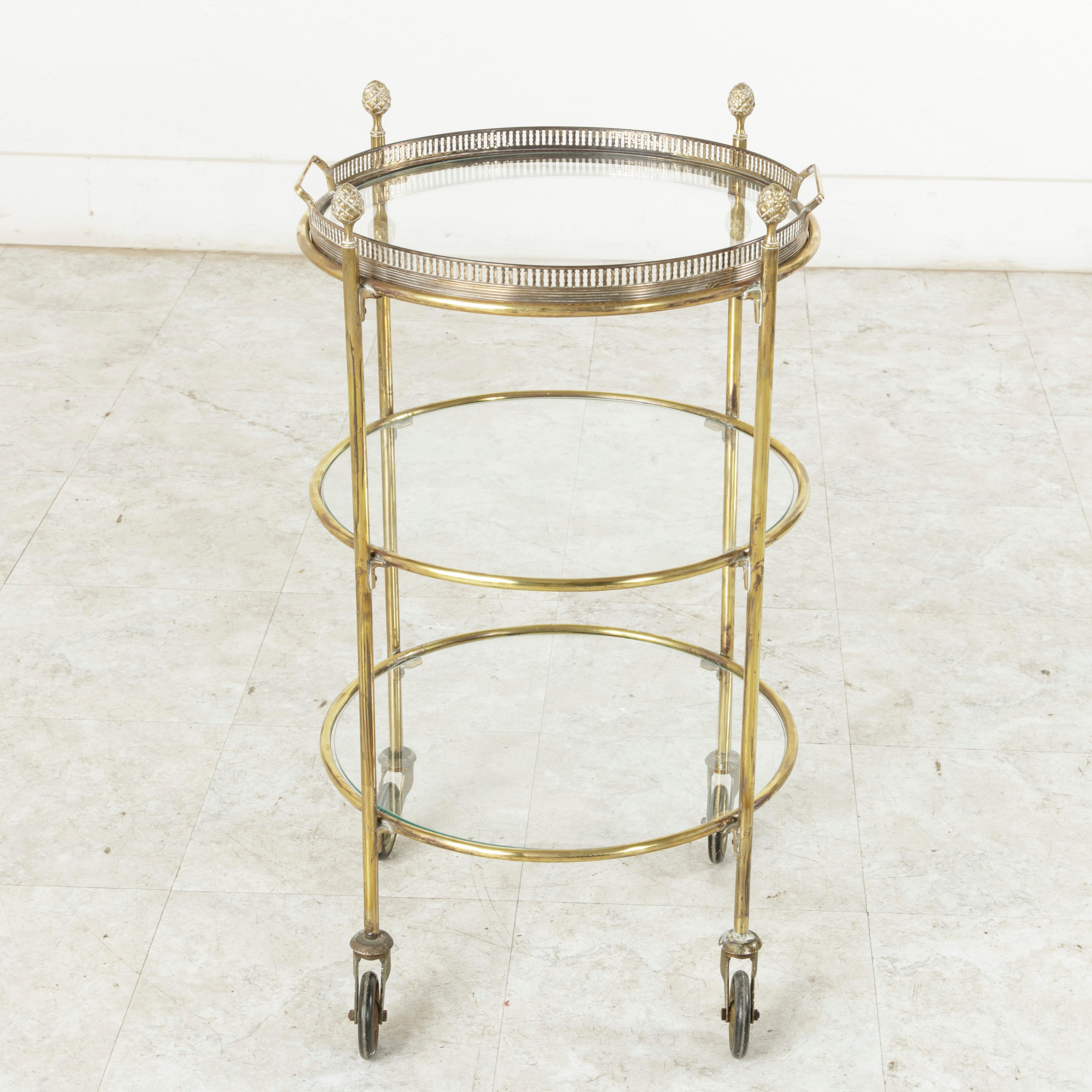 This midcentury French round brass bar cart features three glass shelves and a removable tray on top, with handles, surrounded by a pierced gallery. Its four legs rest on casters and are finished with Louis XVI style pine cone finials. With only a