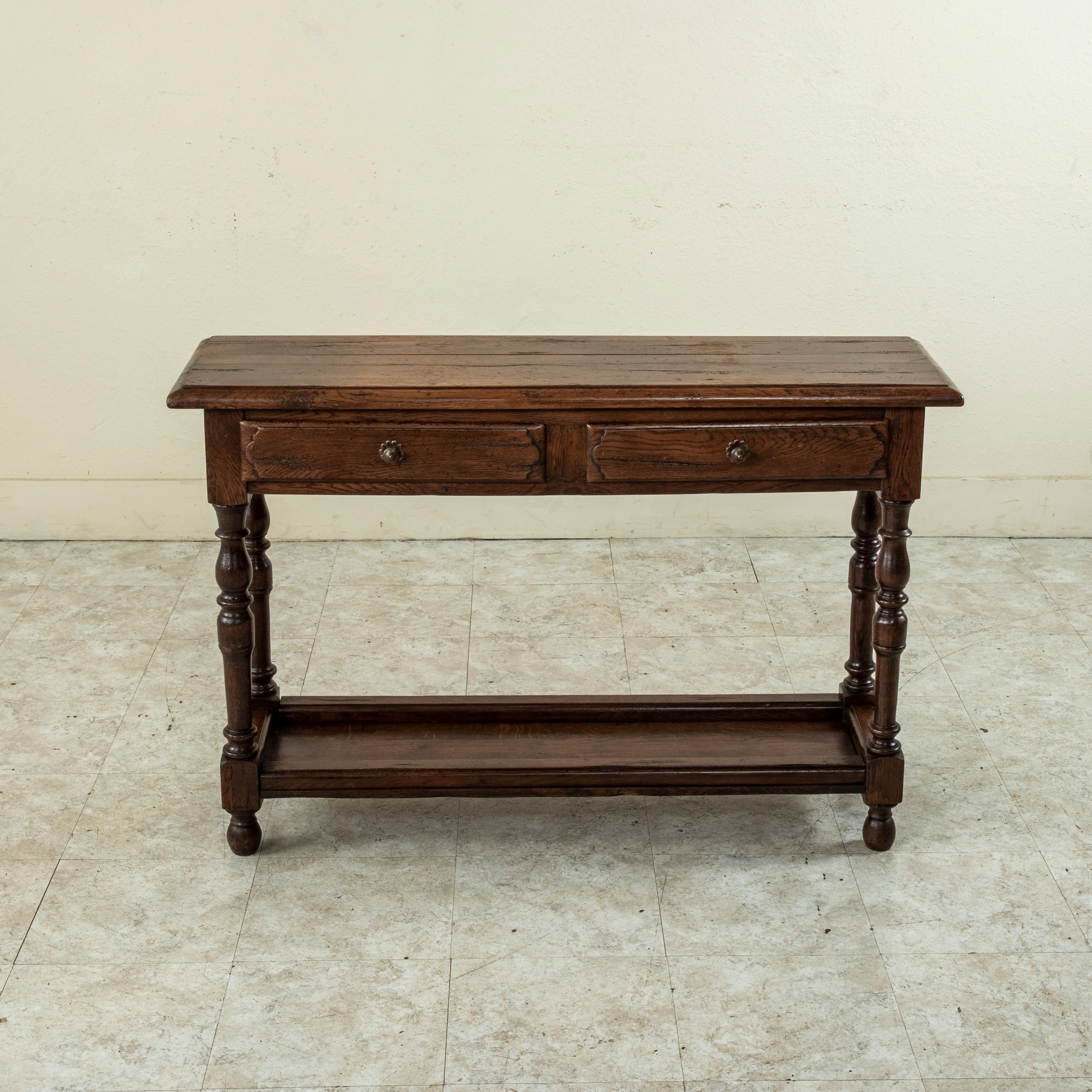 Found in the region of Normandy, France, this Mid-20th Century oak console table features a beveled top and turned legs. Two drawers fit into the apron and are fitted with iron drawer pulls. The legs are joined by a lower shelf that provides