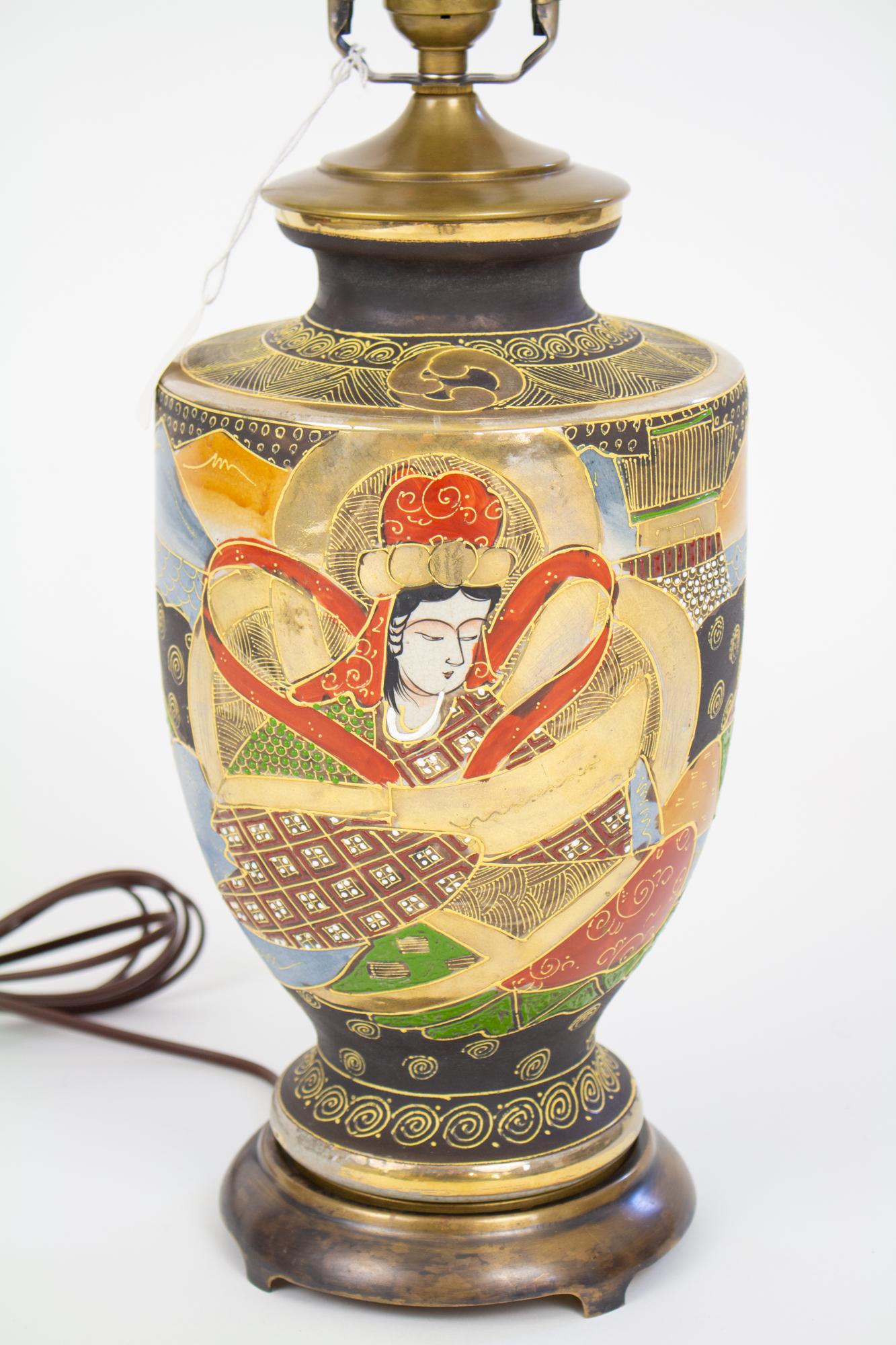 Mid 20th century satsuma table lamp. Features a woman and man in ornate traditional robes, with mountains ocean and a village in the background. Matte black base with red, green and blue details and raised gold accents. Includes harp. Condition: